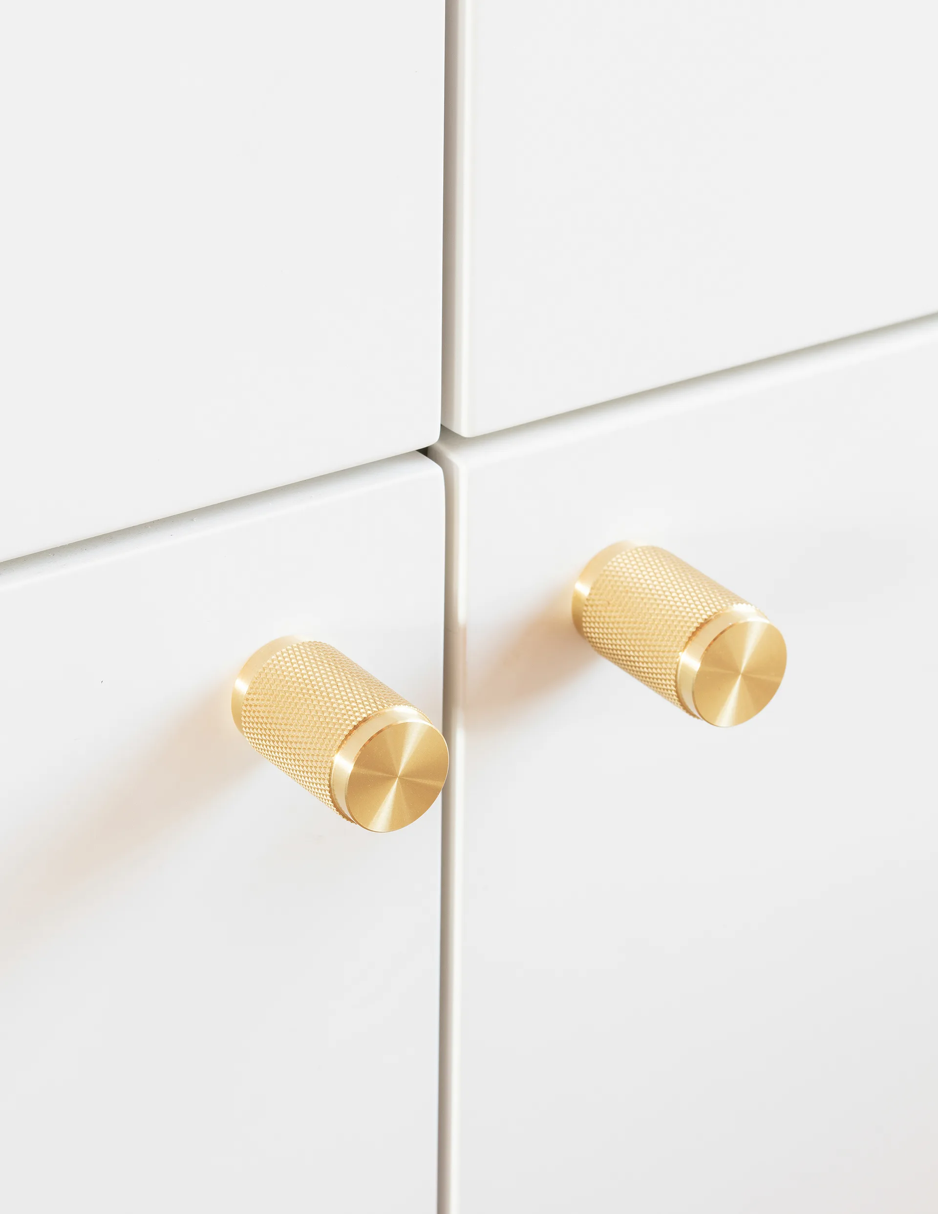 Close-up of two textured cylindrical brass knobs mounted on minimalist flat-panel white cabinet doors.