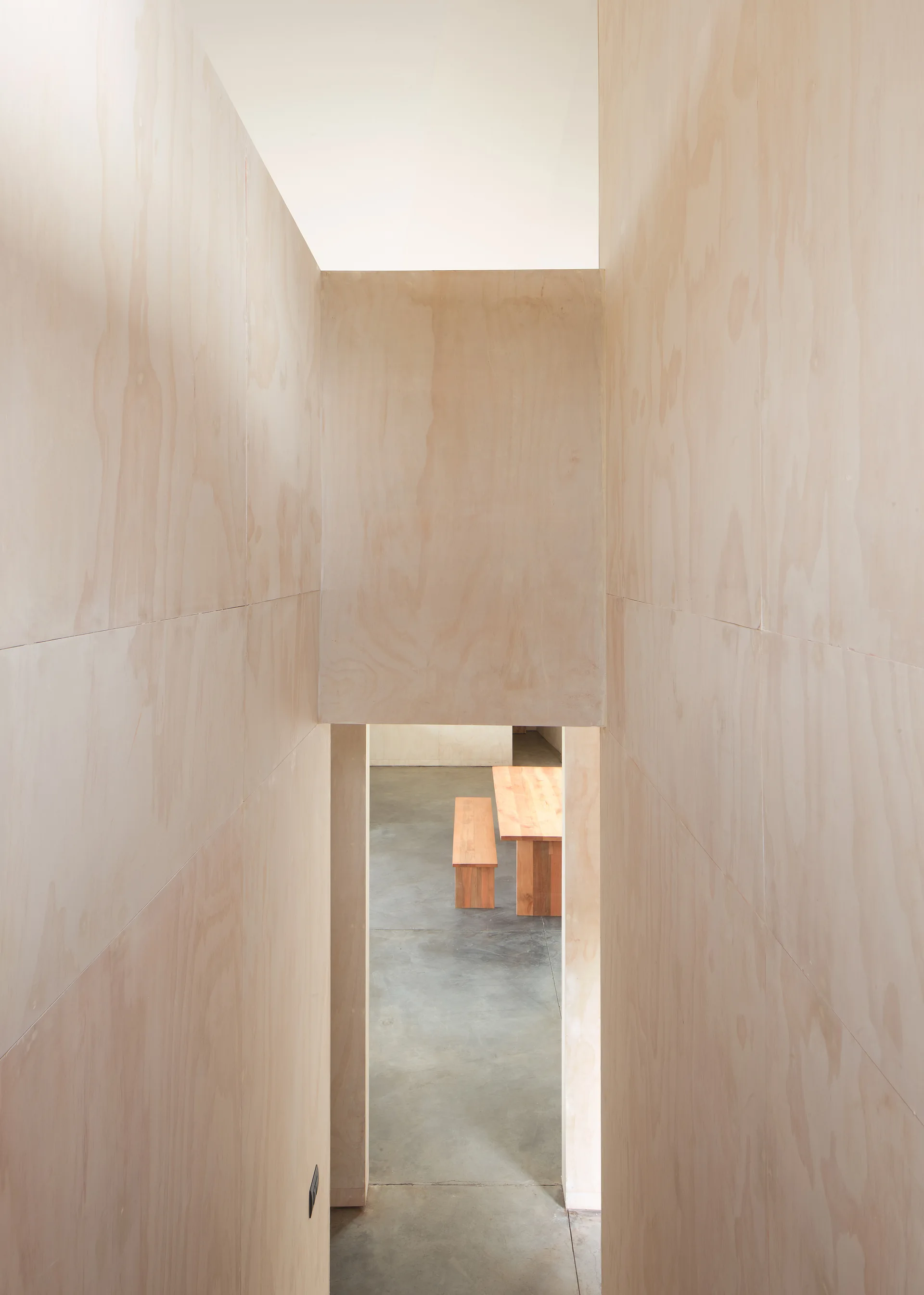 A view looking down a narrow plywood-walled staircase toward a grey concrete floor with a light wood dining table and bench.
