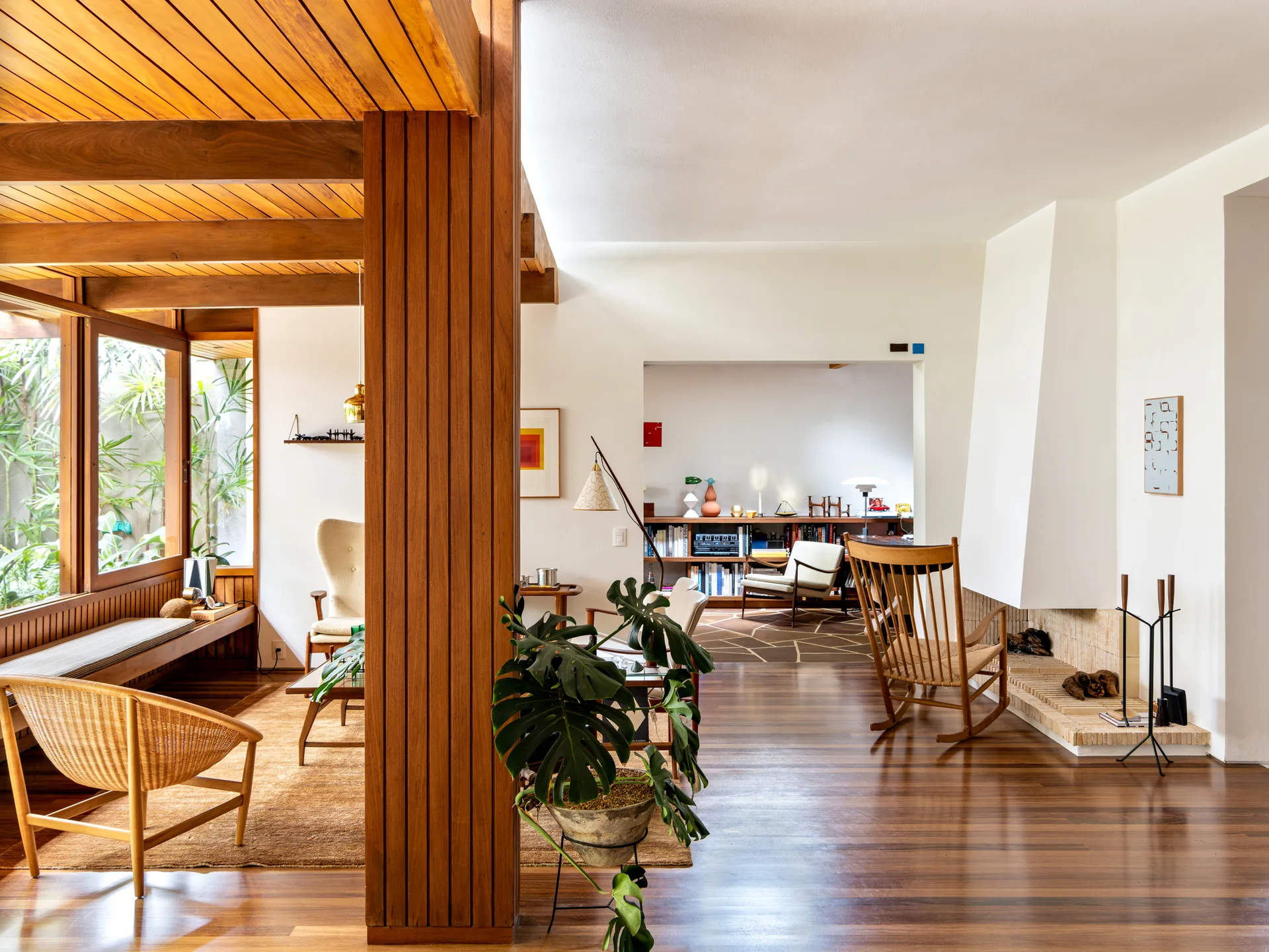 A mid-century modern living room featuring warm wood ceiling beams, a vertical wooden pillar, polished floors, and a minimalist white fireplace.