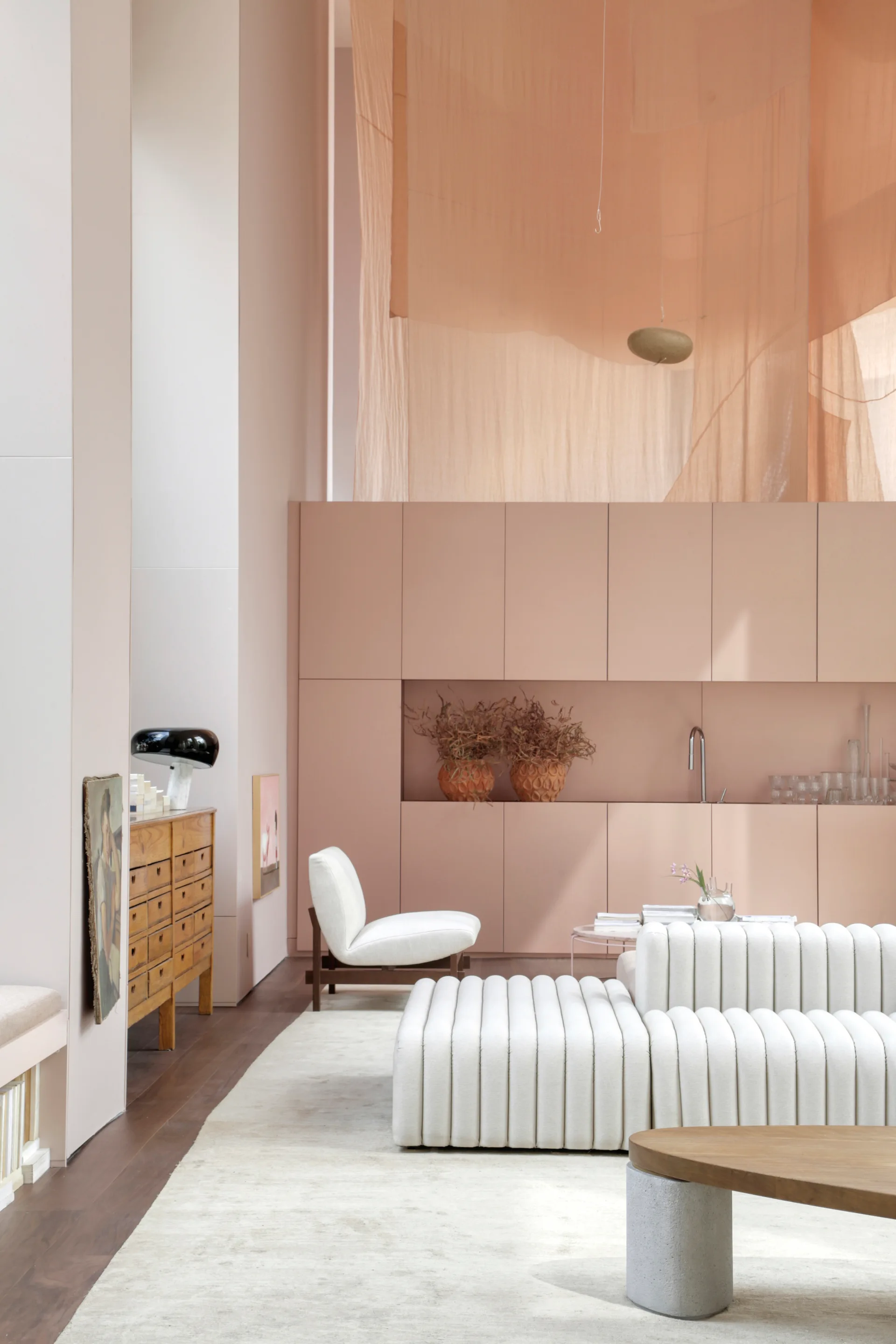 A minimalist living room features a white ribbed sofa, a built-in blush pink storage wall, and draped peach fabric hanging from the high ceiling.