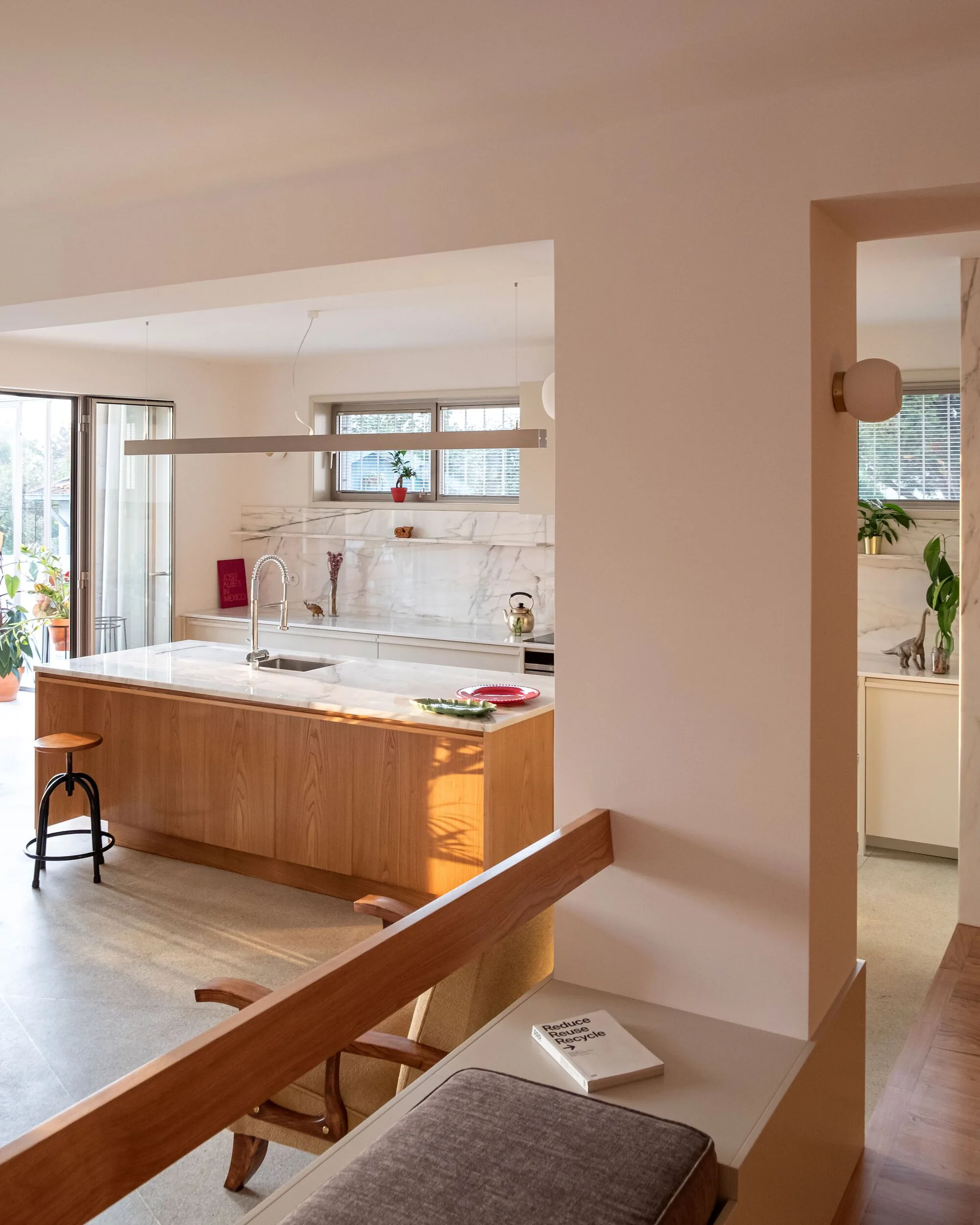 A modern kitchen with a wood island, white marble countertops, and a linear pendant light, viewed through an off-white wall opening.