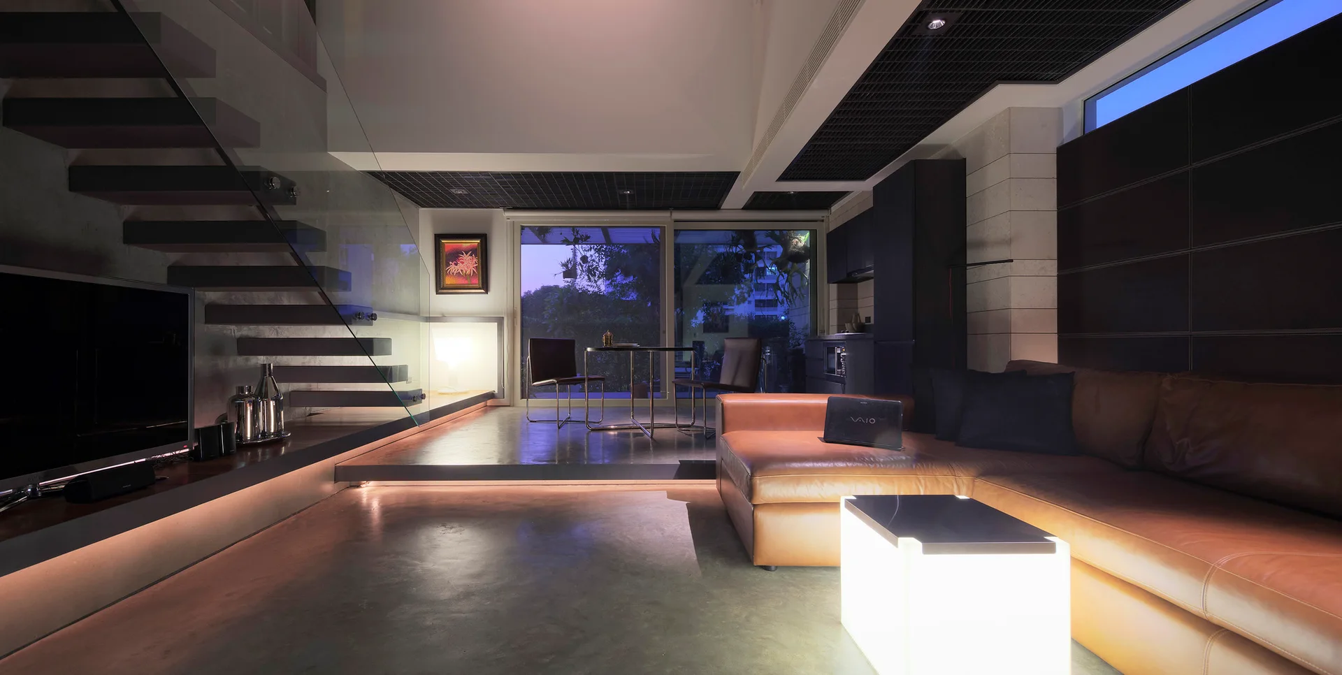 A modern living room features a tan leather sectional sofa, a glowing cube accent table, polished concrete flooring, and a floating staircase.