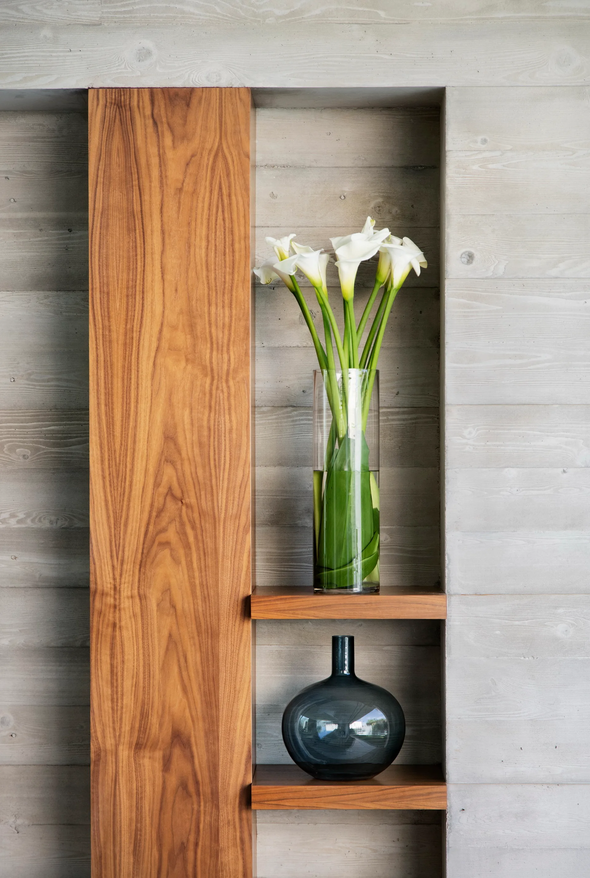 A recessed wall niche in a textured grey concrete wall features warm wood shelving holding white calla lilies in a glass vase and a dark blue vessel.
