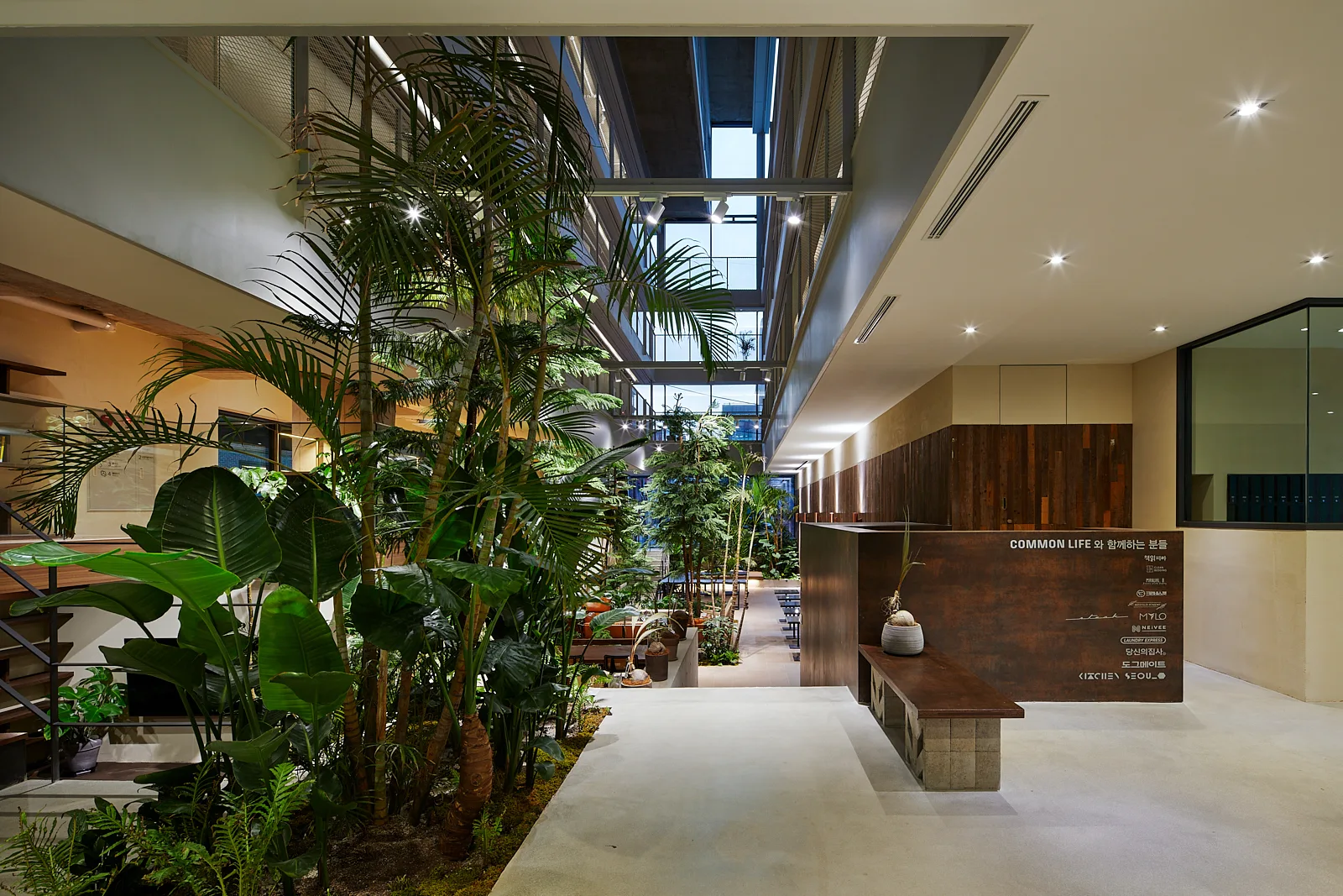 A modern multi-story atrium lobby features a dense cluster of tropical indoor plants, wood-paneled walls, and a concrete floor.