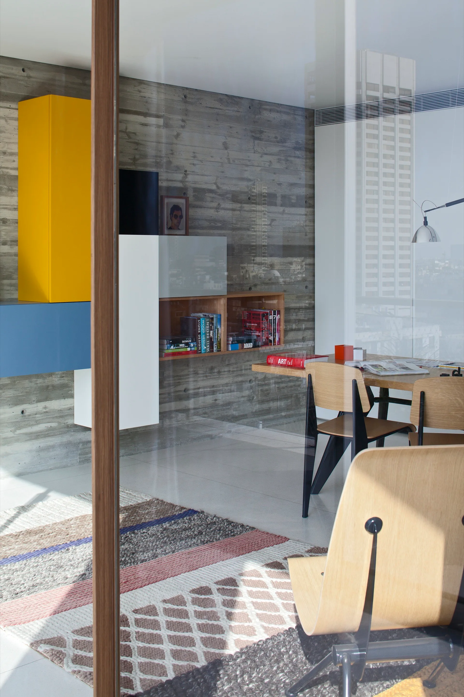 A modern home office with a wood desk and chairs set against a textured concrete wall with yellow and blue floating storage cabinets.