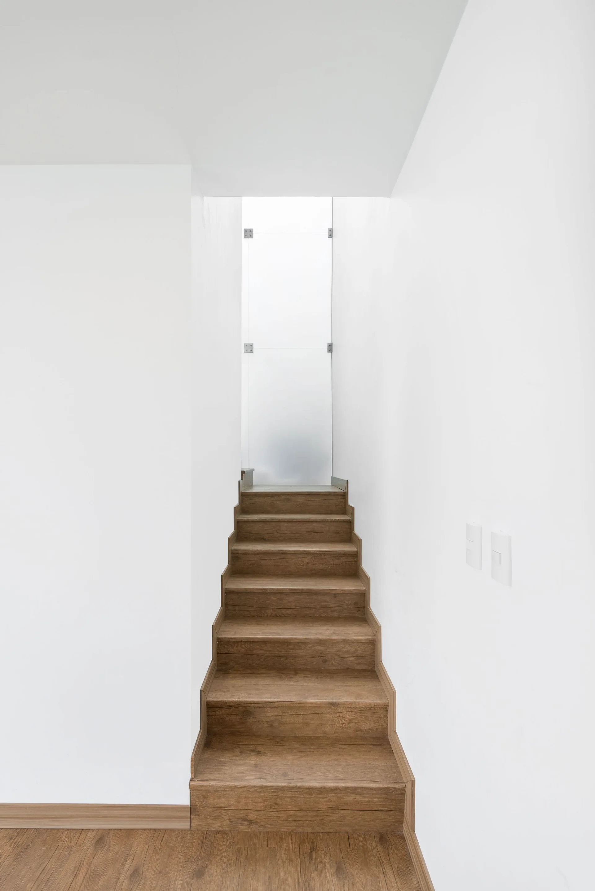 A narrow modern staircase with dark wood-toned treads climbs between solid white walls toward a frosted glass door.