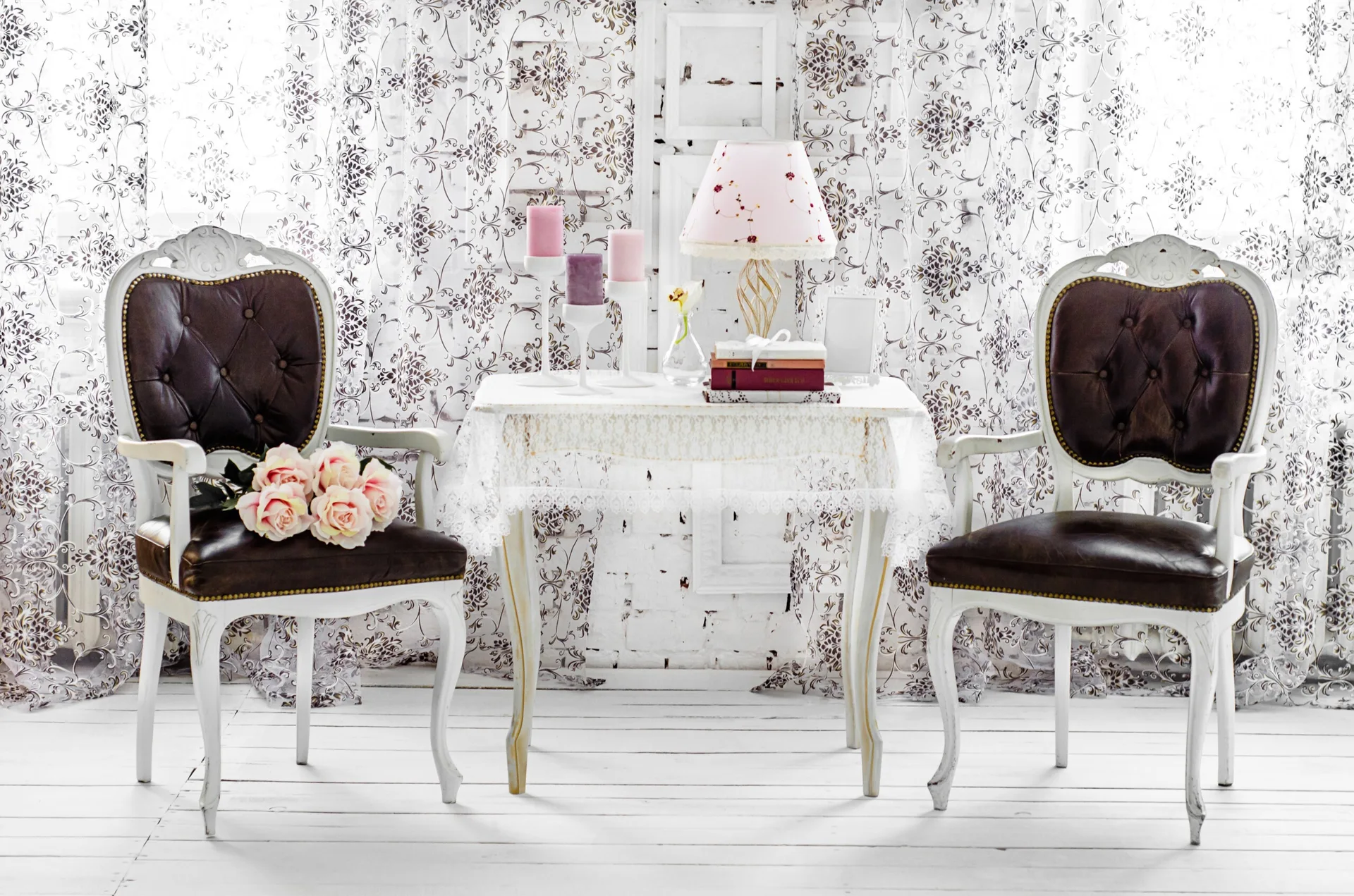 Two vintage white wooden armchairs with dark cushions sit beside a lace-covered table against a floral patterned curtain background.