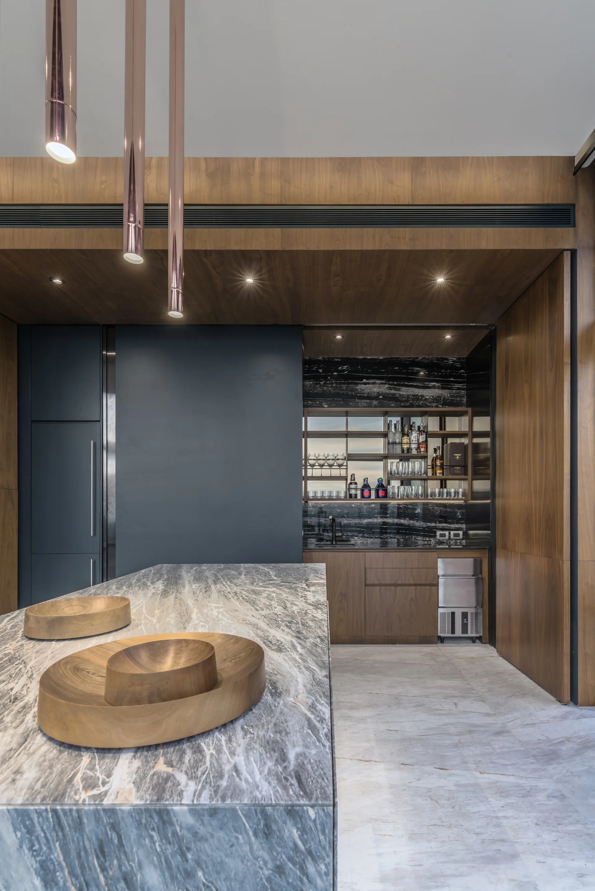 A home bar with warm wood paneling, a marble island with wooden bowls, cylindrical copper pendant lights, and a recessed wet bar with a dark backsplash.
