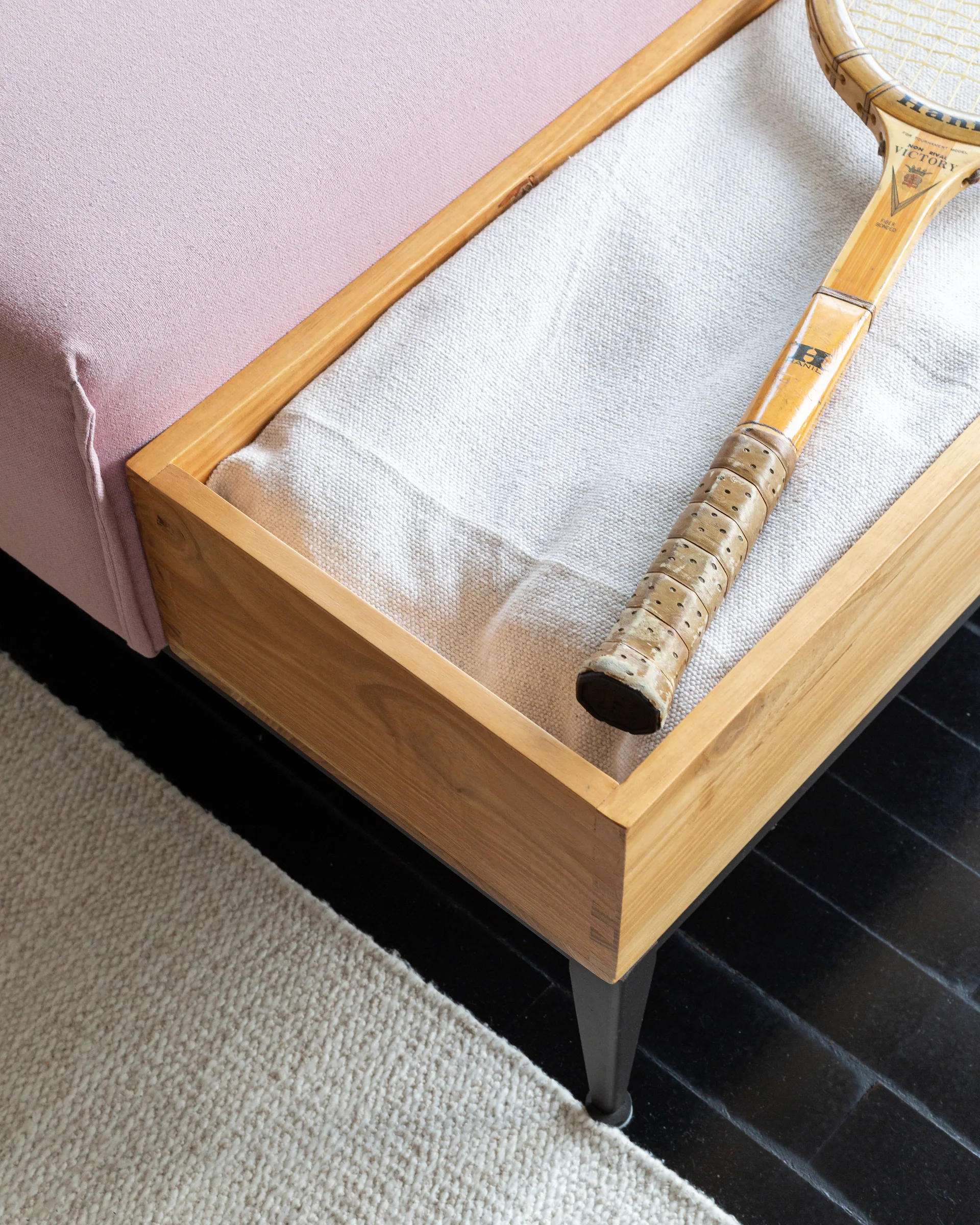 An upholstered pink bench features an inset wooden tray holding a cream textile throw and a vintage wooden tennis racket on a dark floor.