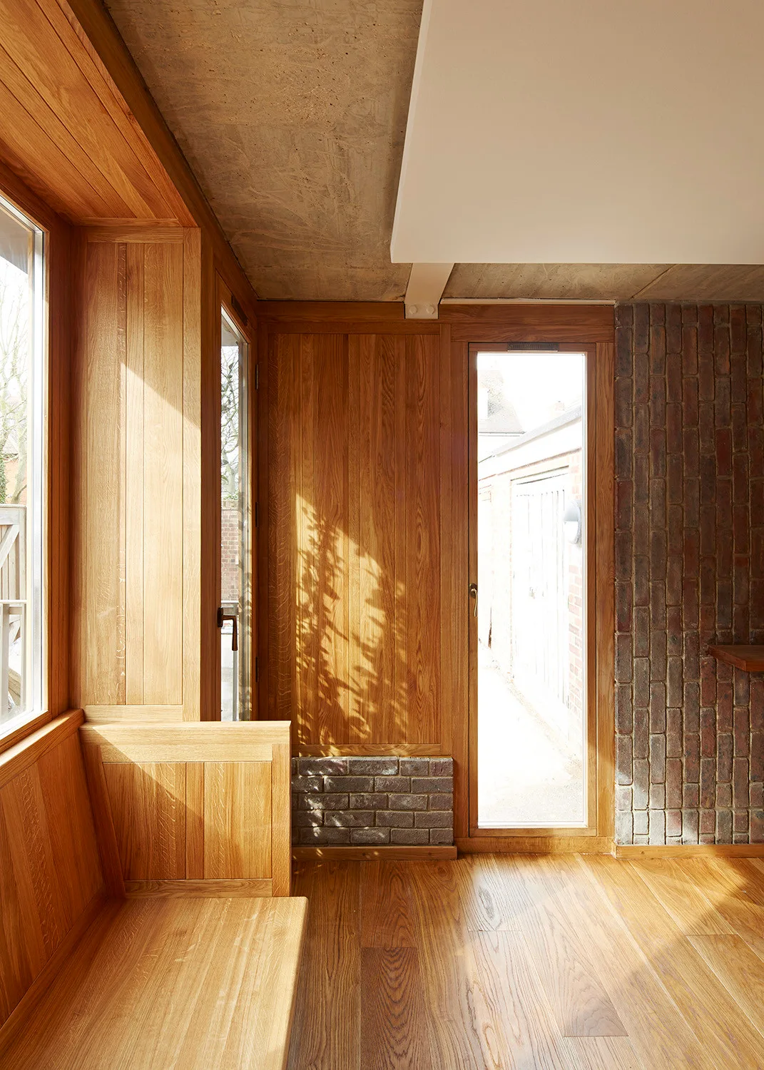 A sunlit entryway featuring natural wood wall paneling, a matching built-in bench, a raw concrete ceiling, and an exposed brick wall.