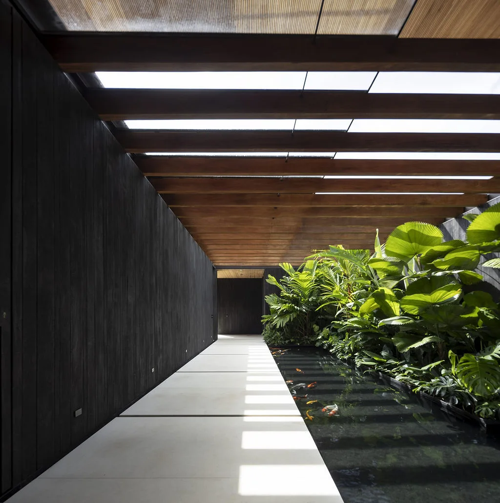 A light paved walkway runs alongside a long koi pond with green plants under a slatted wooden ceiling and next to a dark wood wall.
