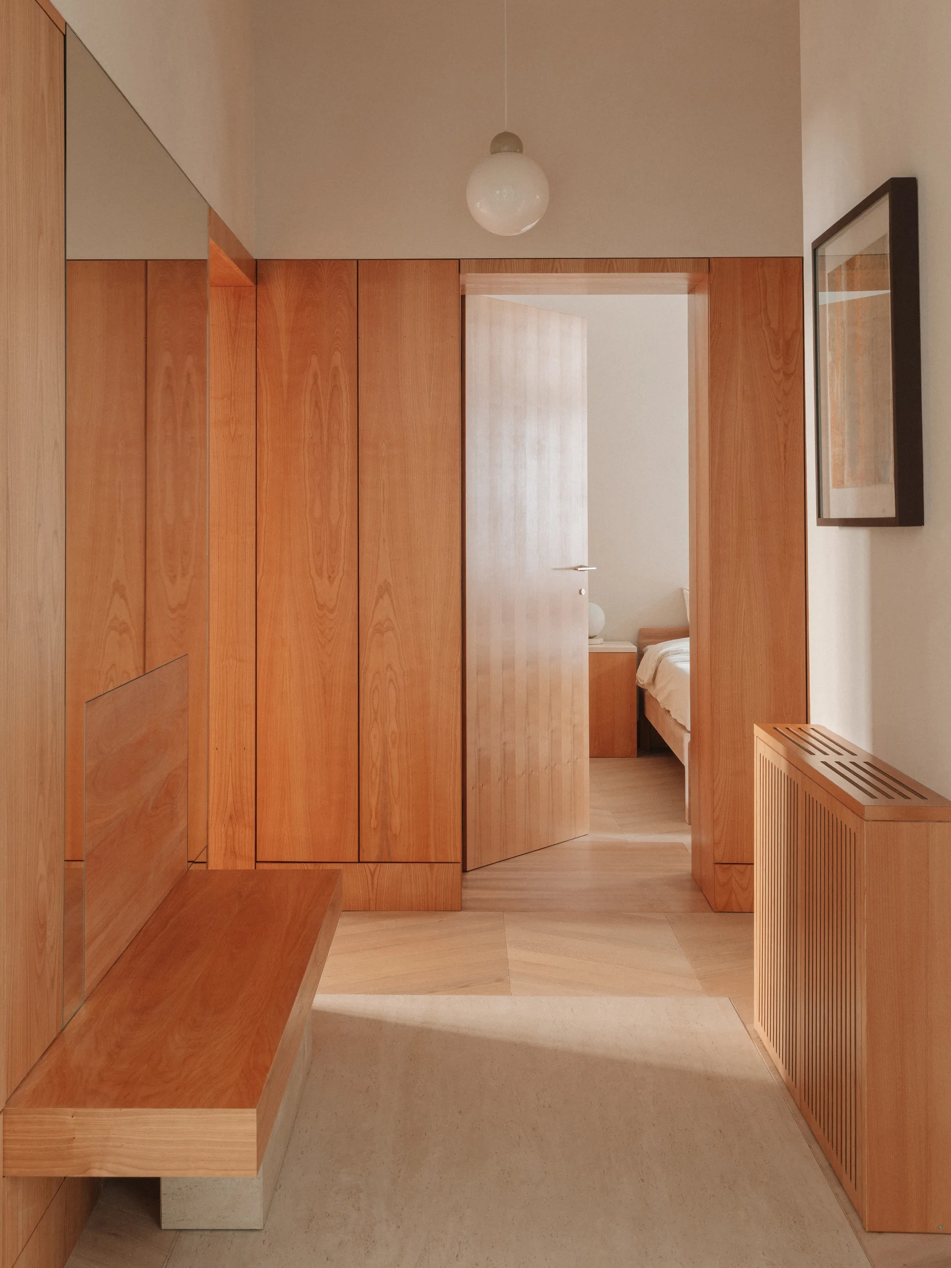 A minimalist hallway features honey-toned wood wall panels, a matching floating bench, a slatted radiator cover, and an open doorway leading to a bedroom.