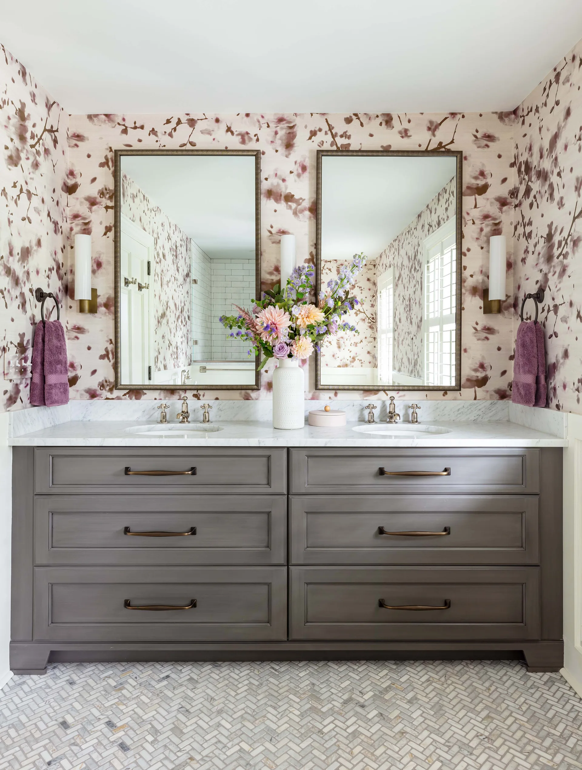 A bathroom features a grey double vanity with a marble countertop, two mirrors, purple floral wallpaper, and a herringbone tile floor.