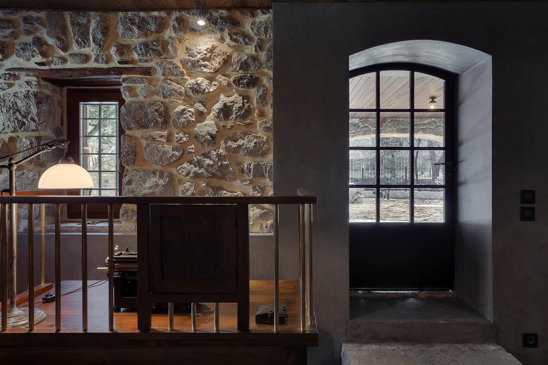 An interior space features a rough exposed stone wall with a small window, a dark wood railing, and an arched glass door set in a smooth gray wall.