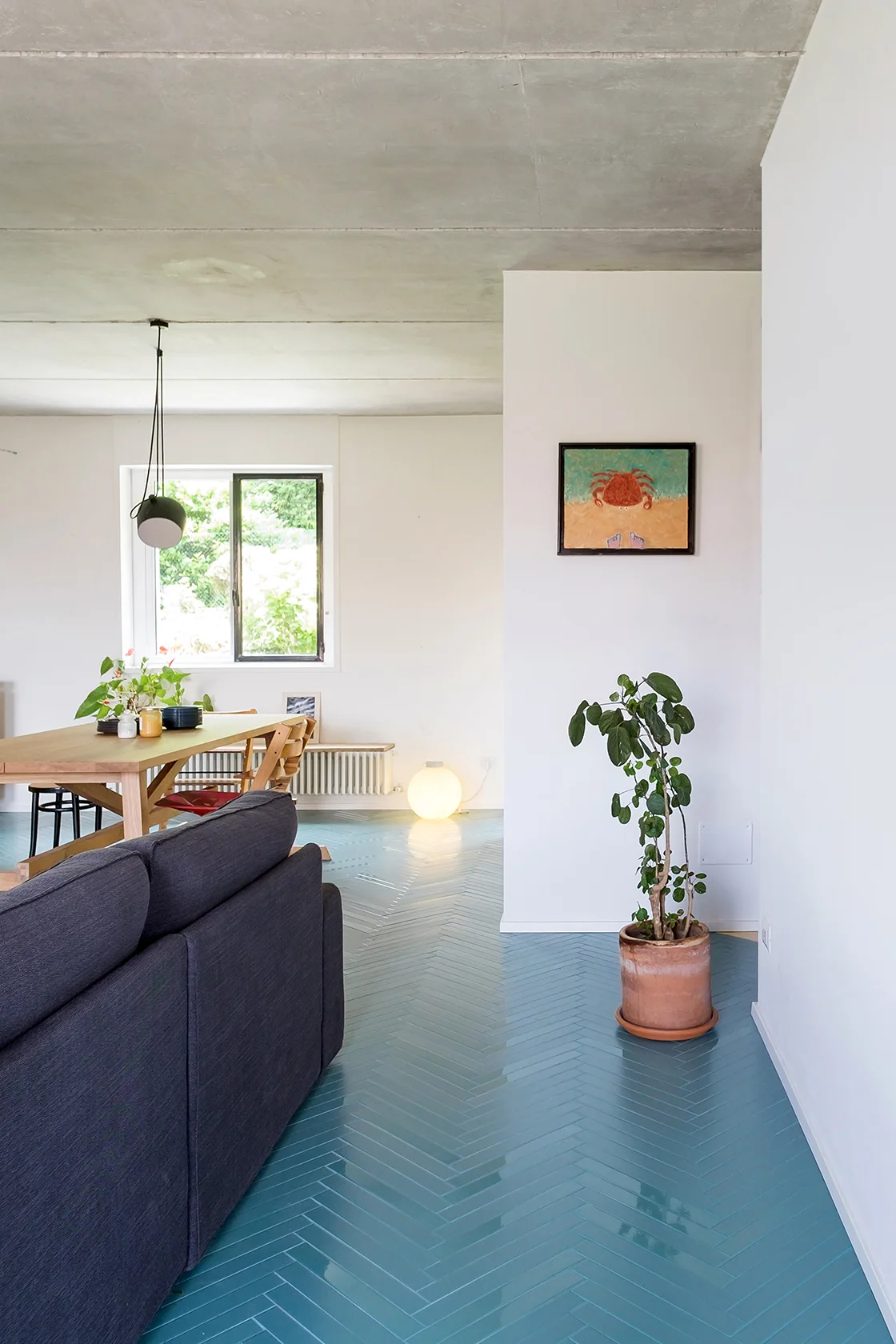 An open-plan living space features a glossy teal herringbone tile floor, a dark grey sofa, a wooden dining table, and an exposed concrete ceiling.