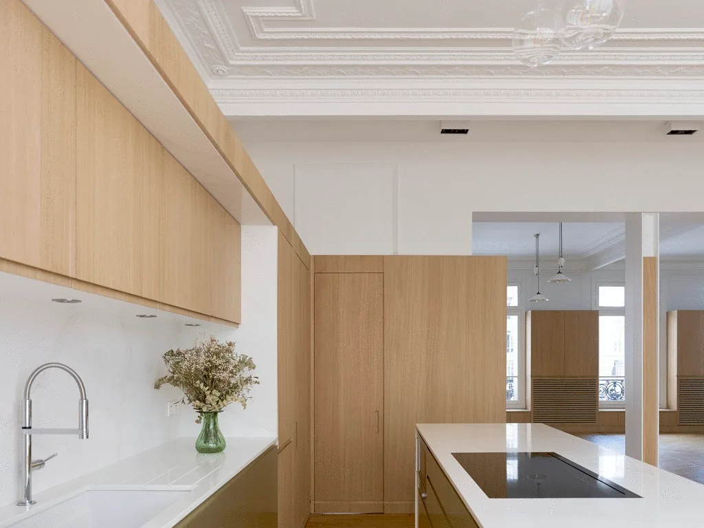 A modern kitchen with light wood cabinetry, white stone countertops, an island with a cooktop, and an ornate white plaster ceiling.