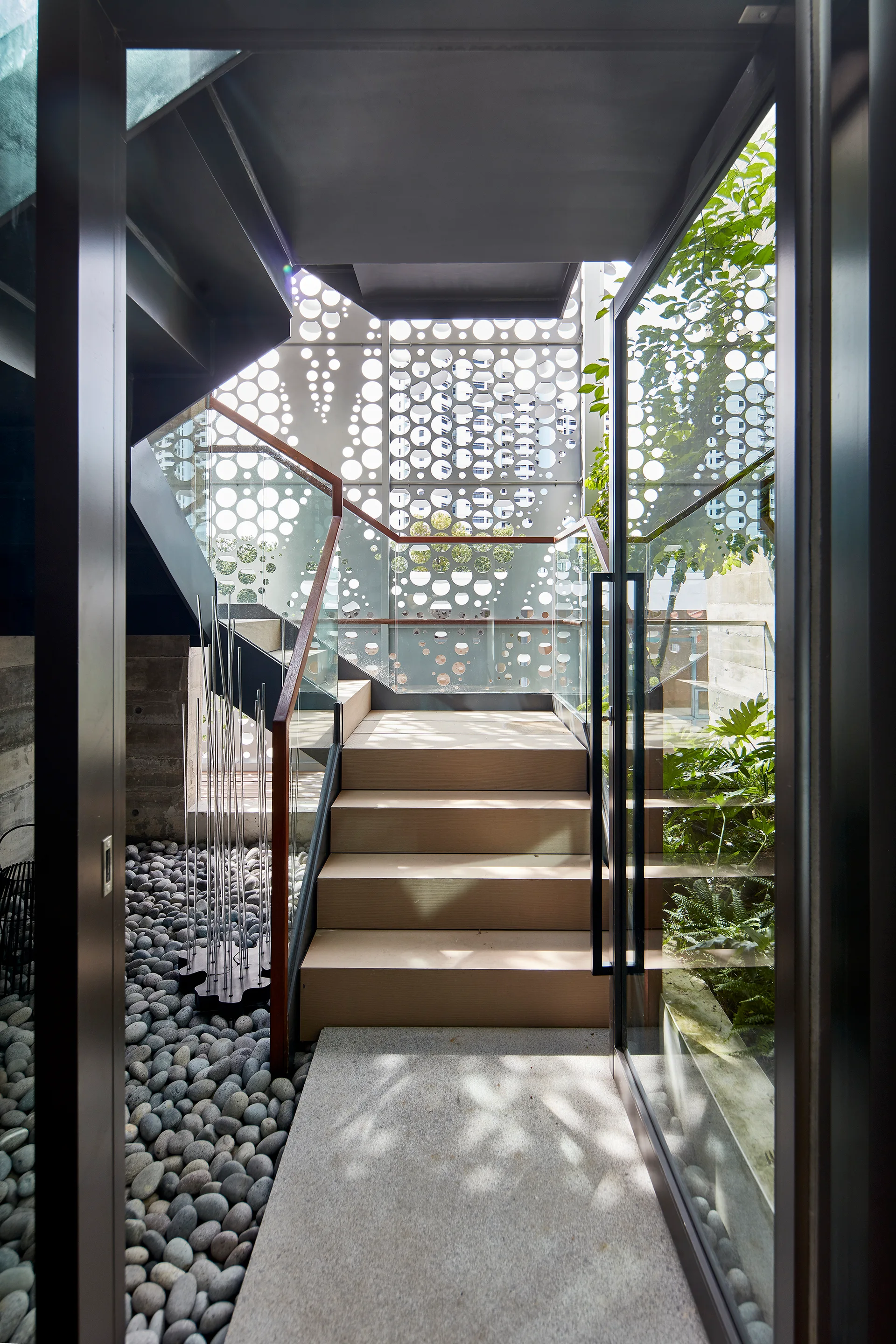 A modern indoor staircase features wooden treads, glass railings, and a large perforated metal screen wall above a bed of grey river pebbles.
