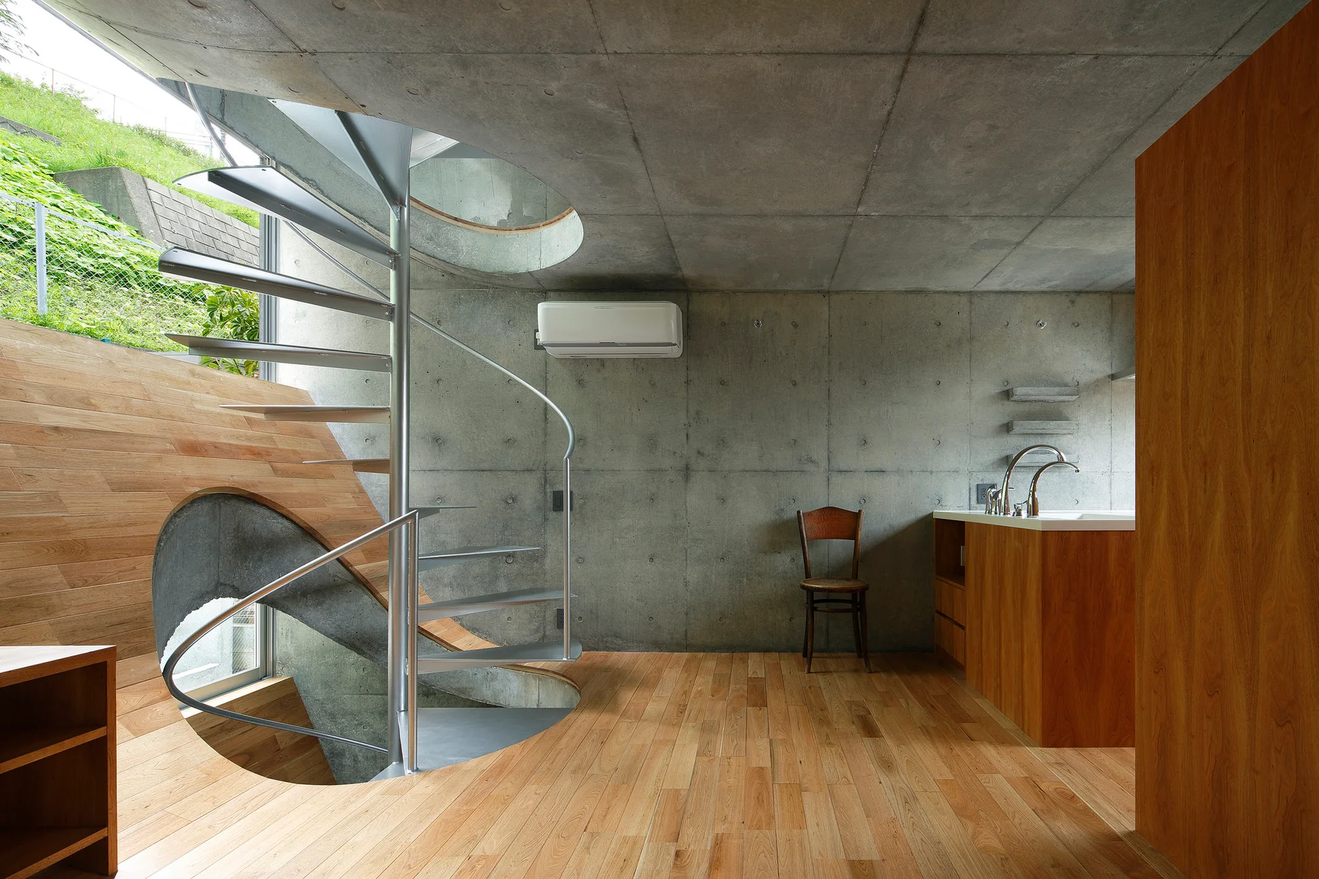 A metal spiral staircase rises through a circular opening in a minimalist kitchen with exposed concrete walls, light wood flooring, and a wooden island.