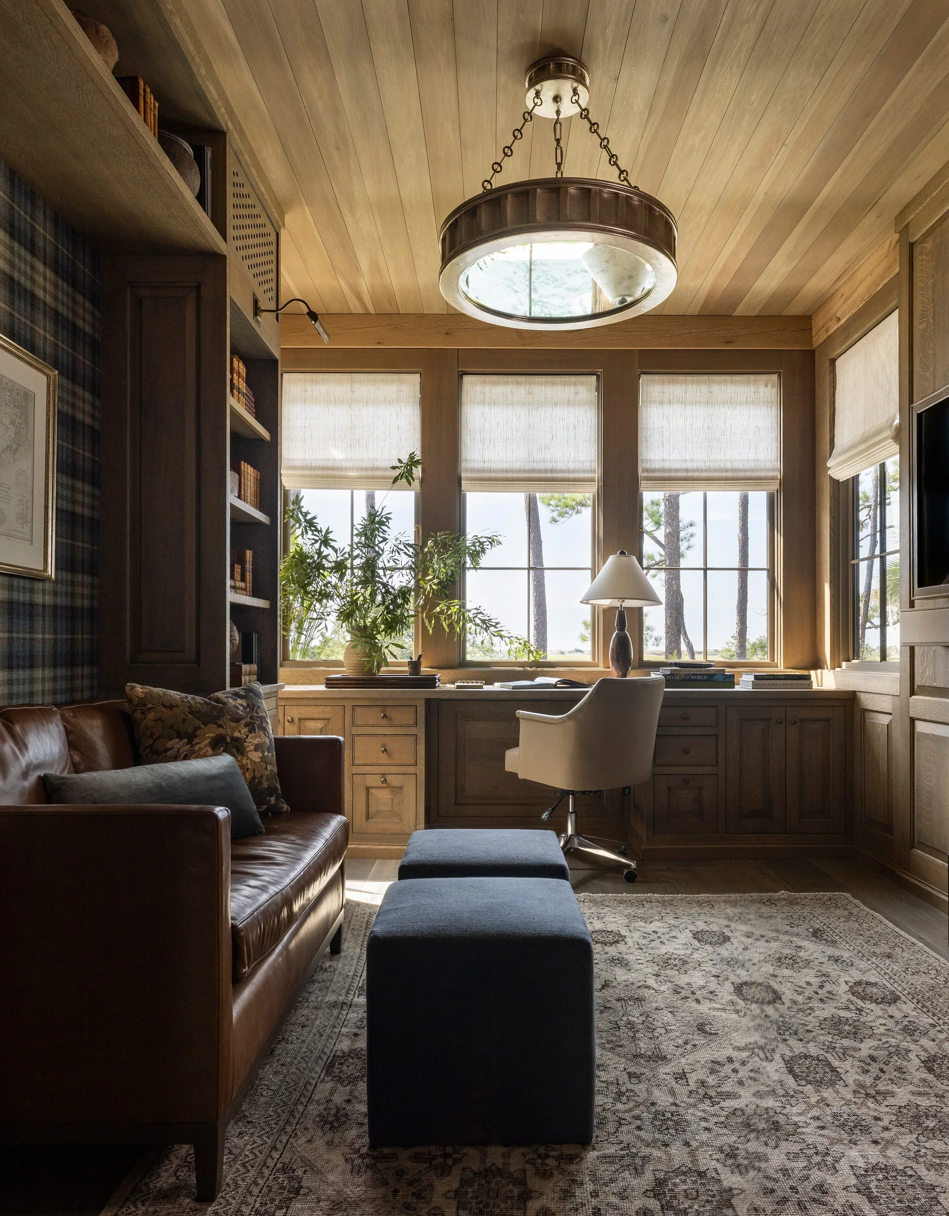 A wood-paneled home office features a built-in desk beneath windows, a brown leather sofa, and two dark blue ottomans on a patterned rug.