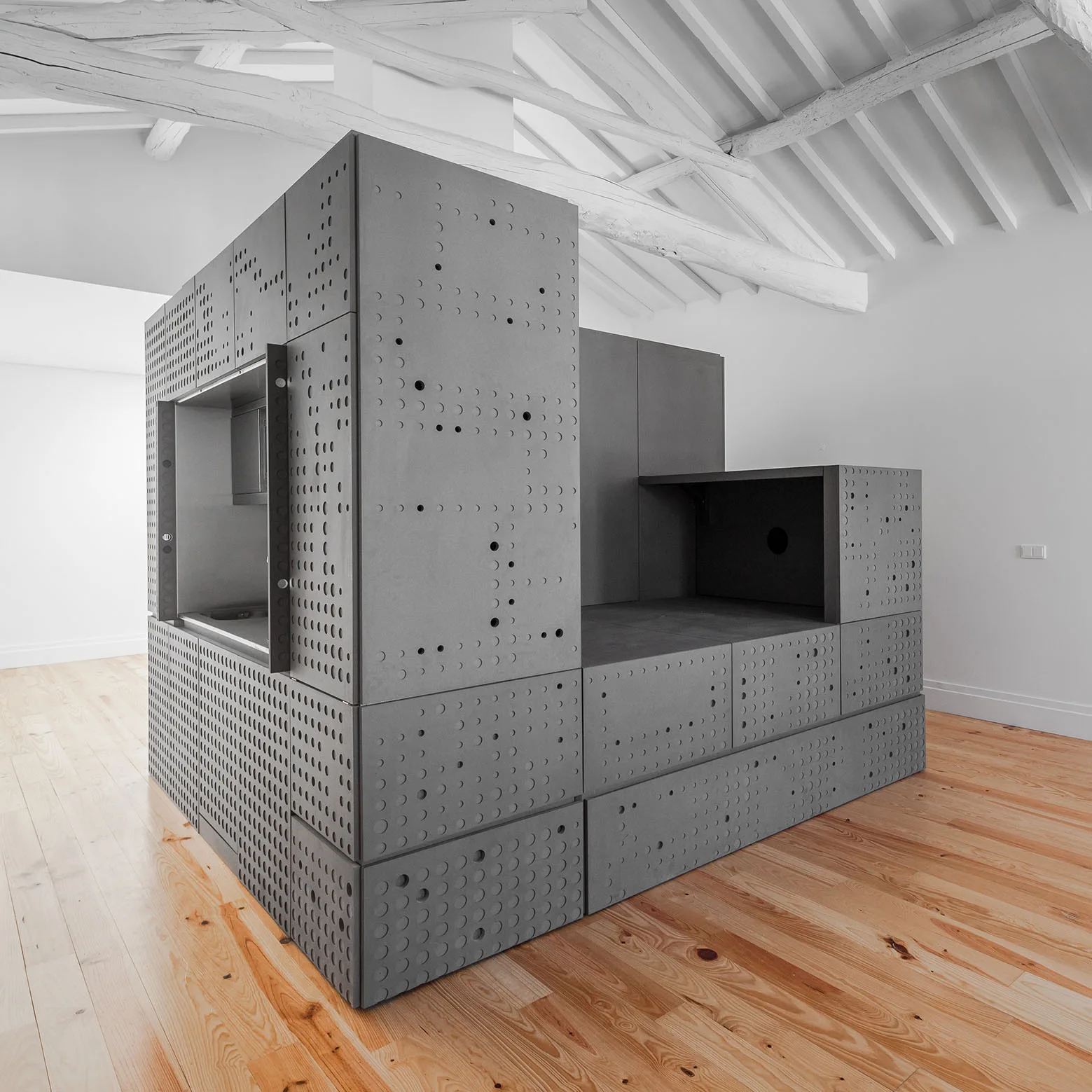 A monolithic gray perforated storage unit sits on a light wood plank floor in a white room with exposed timber ceiling beams.