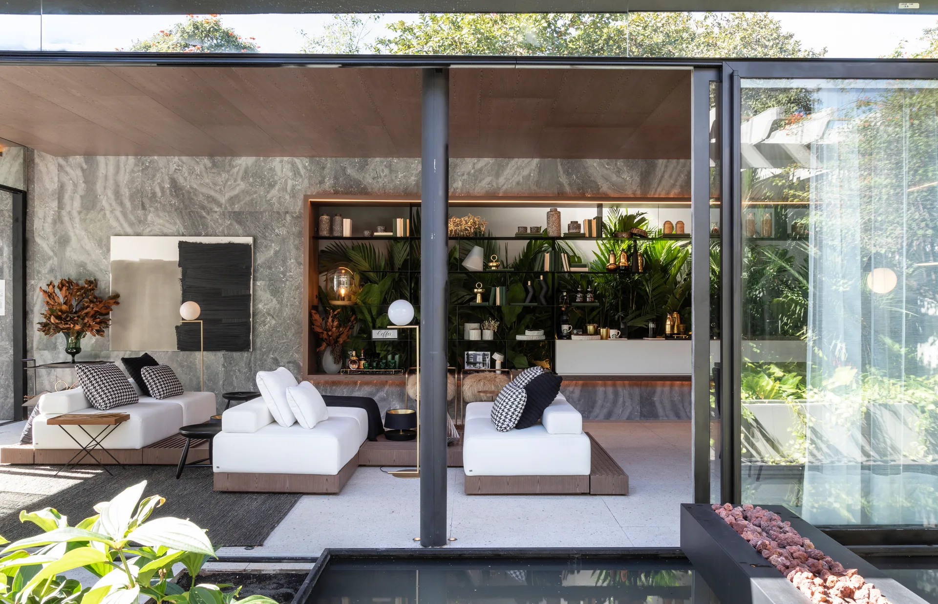 A contemporary indoor-outdoor living room features low-profile white sofas, grey veined stone walls, and a large backlit shelving unit filled with green plants.