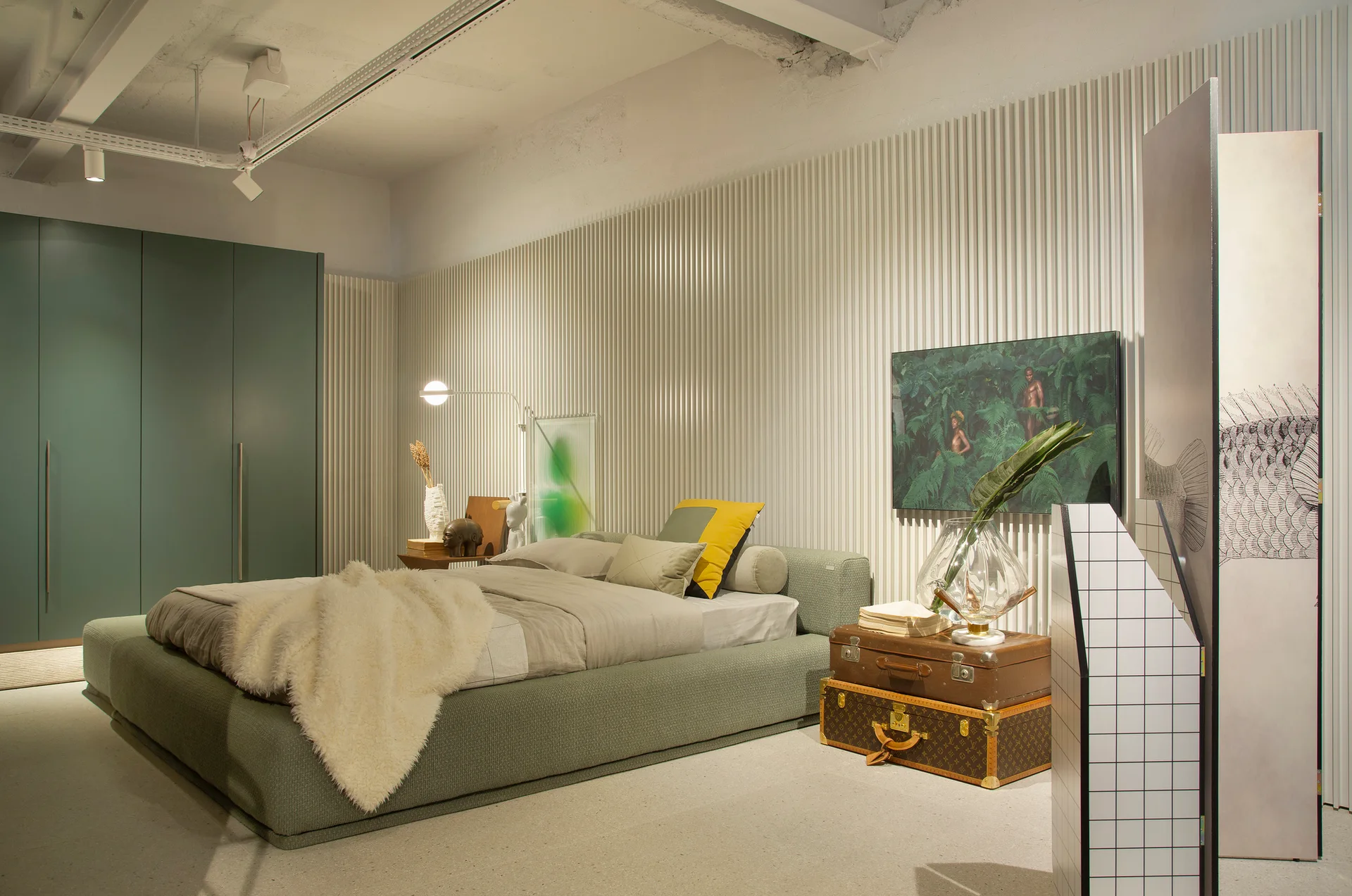A modern bedroom features an upholstered sage green bed, a vertical ribbed wall, and stacked vintage trunks used as a bedside table.
