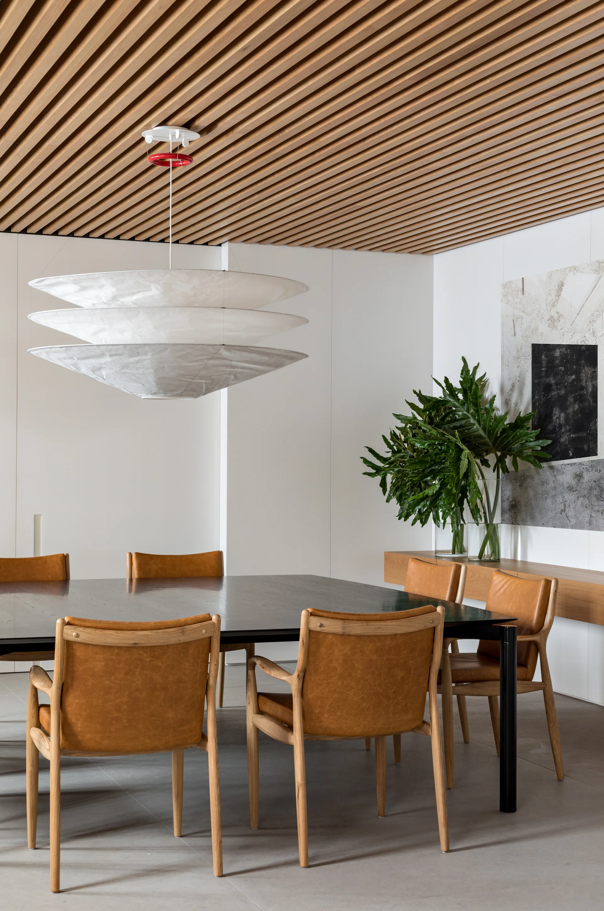 A modern dining room features a wood slatted ceiling, a large white pendant light, and tan leather chairs around a black table.
