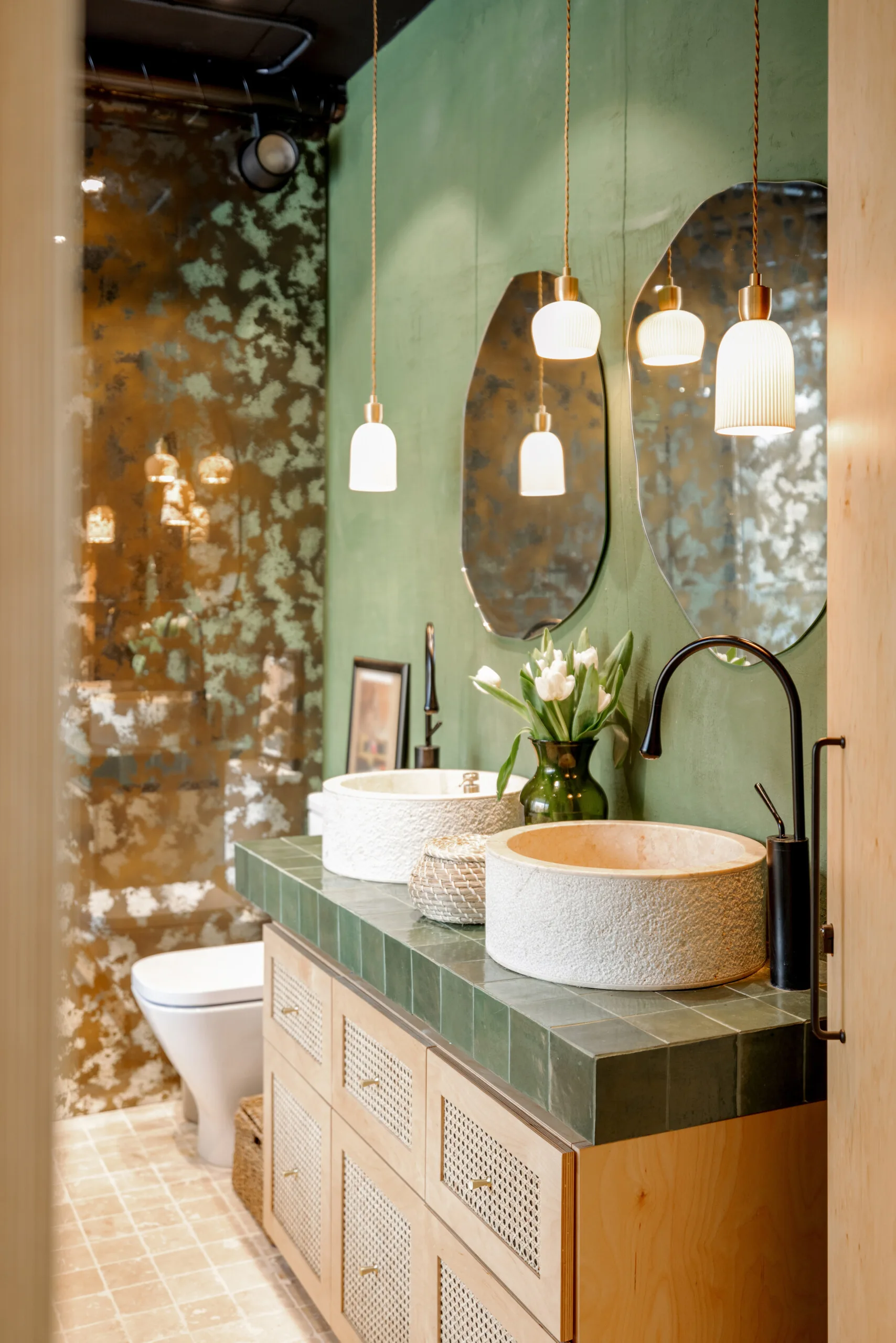A bathroom vanity features two light-colored stone vessel sinks, green tile countertops, cane-front cabinets, and oval mirrors.