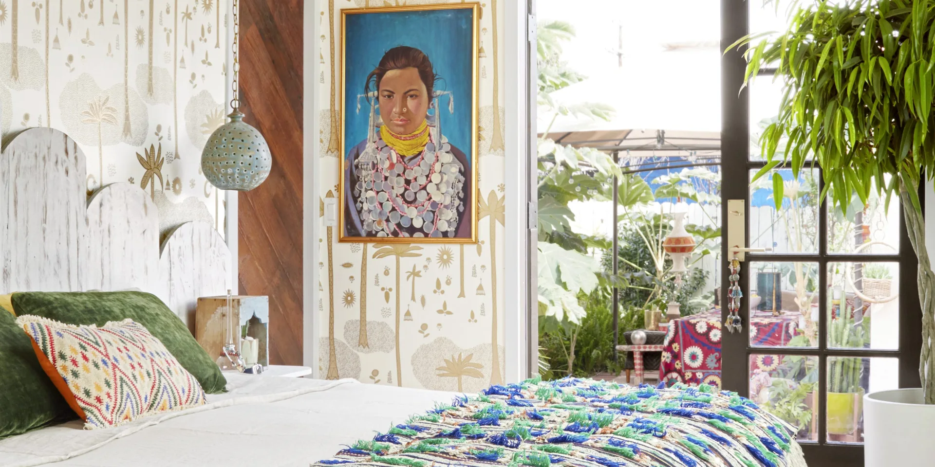 An eclectic bedroom featuring botanical wallpaper, a framed portrait, and a glass door that opens to a lush garden patio.