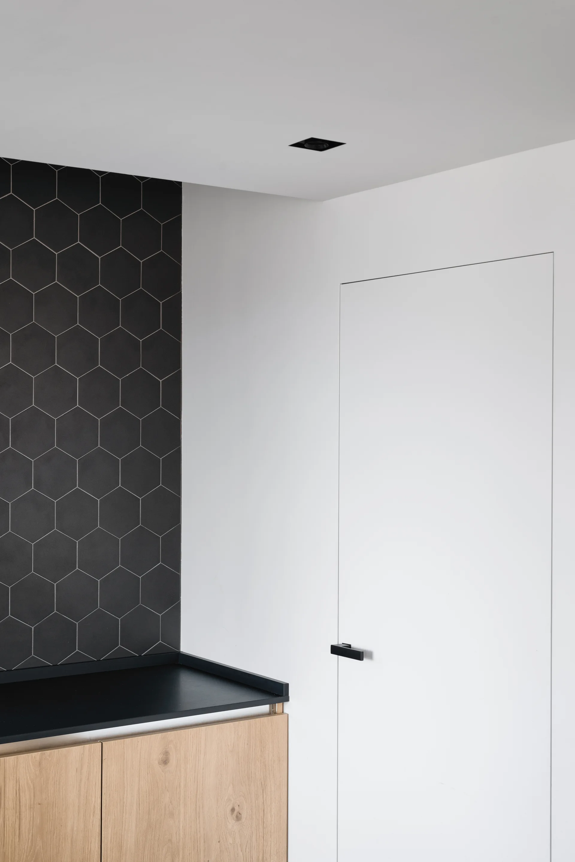 A minimalist room corner featuring a light wood cabinet, a black countertop, black hexagonal wall tiles, and a flush white door with a black handle.