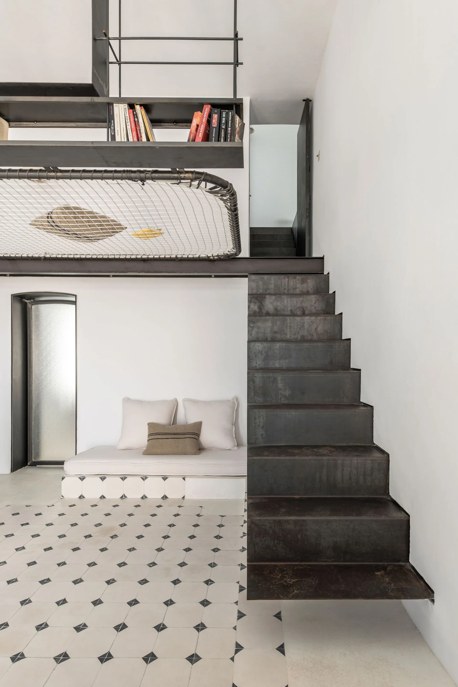 A dark metal staircase leads to a loft mezzanine with a hammock net and shelving above a built-in cushioned daybed and geometric tiled floor.