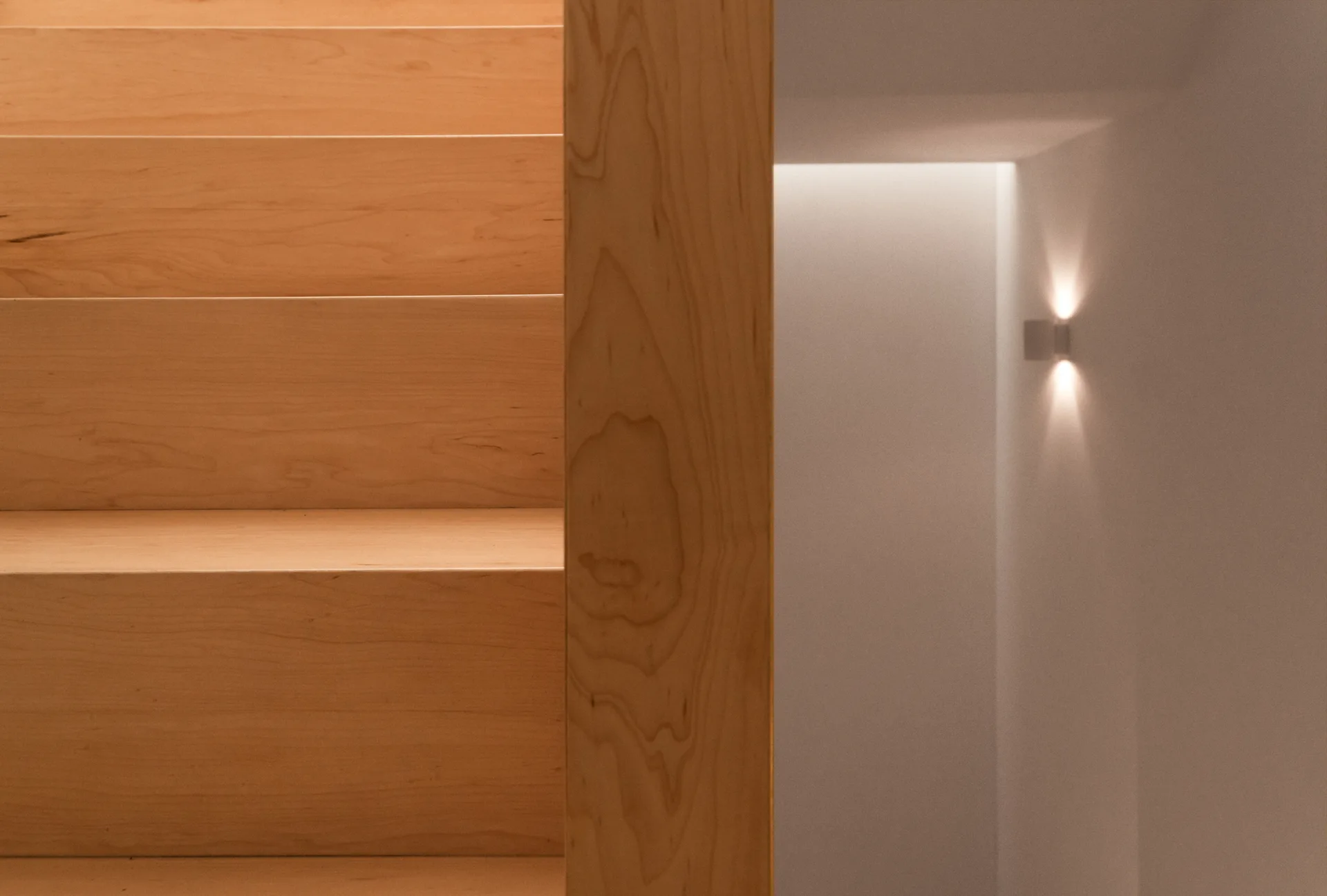 A minimalist light wood staircase next to an off-white plaster wall featuring a small bidirectional wall sconce that casts a warm glow.