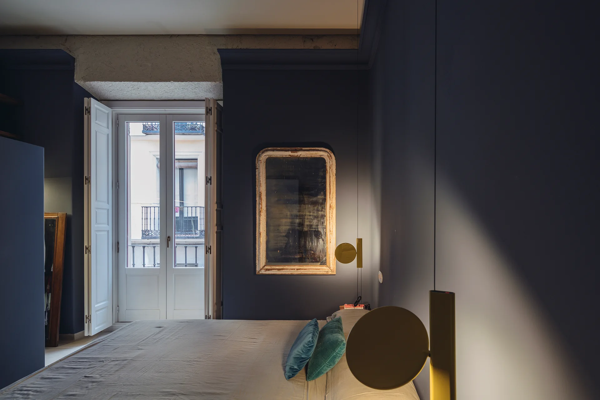 A dark blue bedroom features a bed with blue velvet pillows, an antique wood-framed mirror, and a yellow modern pendant light next to white shuttered doors.