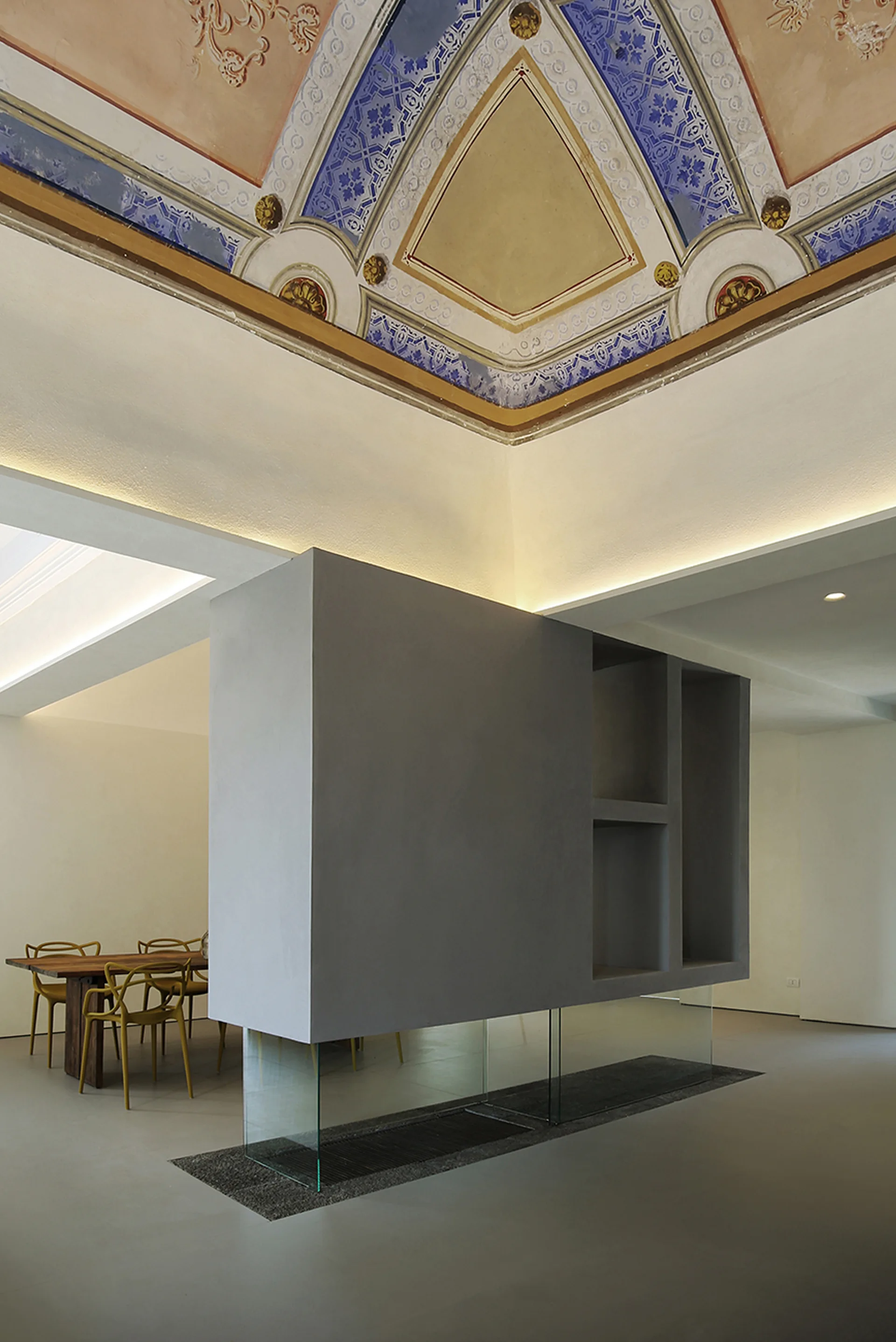 A contemporary gray room divider supported by clear glass panels stands in a dining room beneath a historic frescoed ceiling.