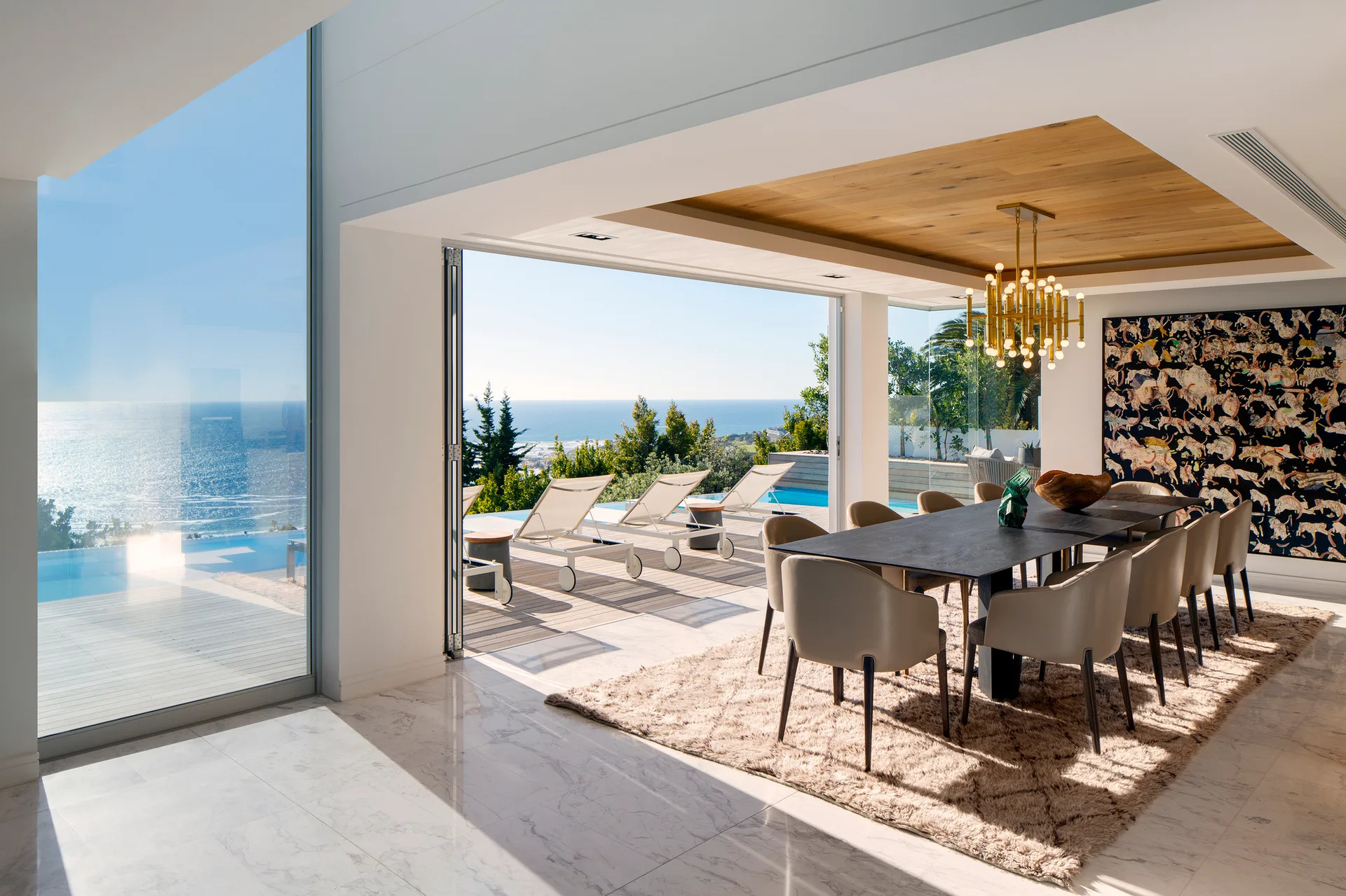 A contemporary dining room features a dark wood table, beige chairs, a brass chandelier, and sliding glass walls that open to a sun deck and ocean.