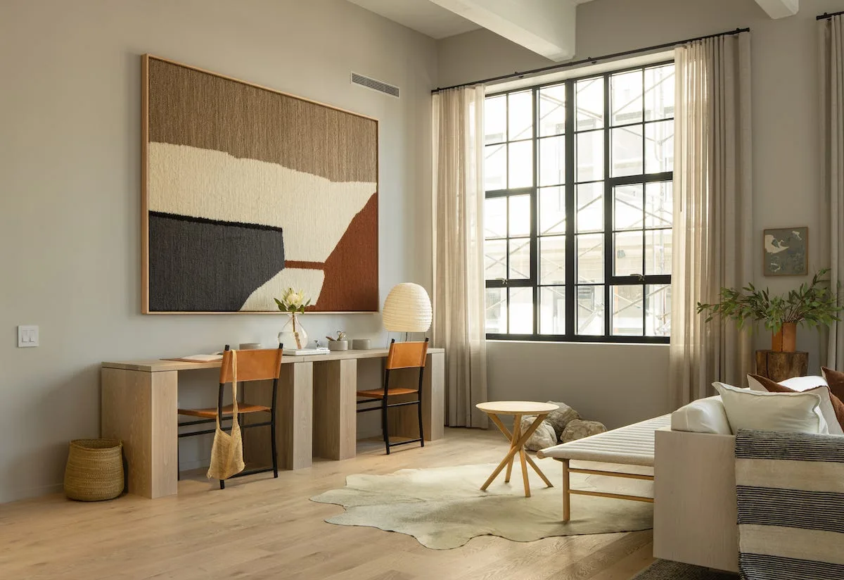A minimalist workspace features a long light-wood desk with two leather chairs, a large woven wall hanging, and a black steel grid window.