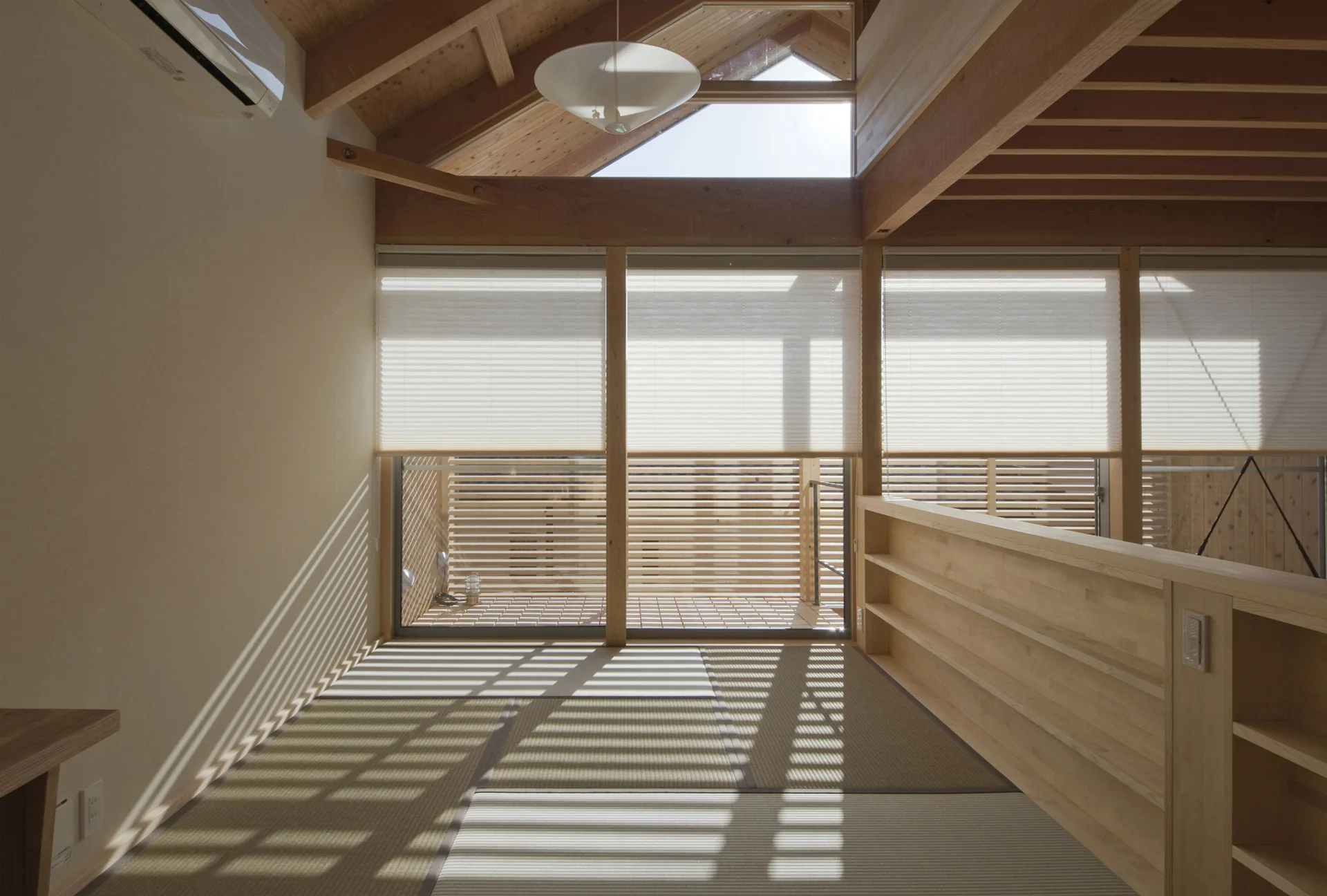 A minimalist loft landing features tatami mat flooring, a low built-in wooden bookcase, and pleated blinds filtering soft daylight.