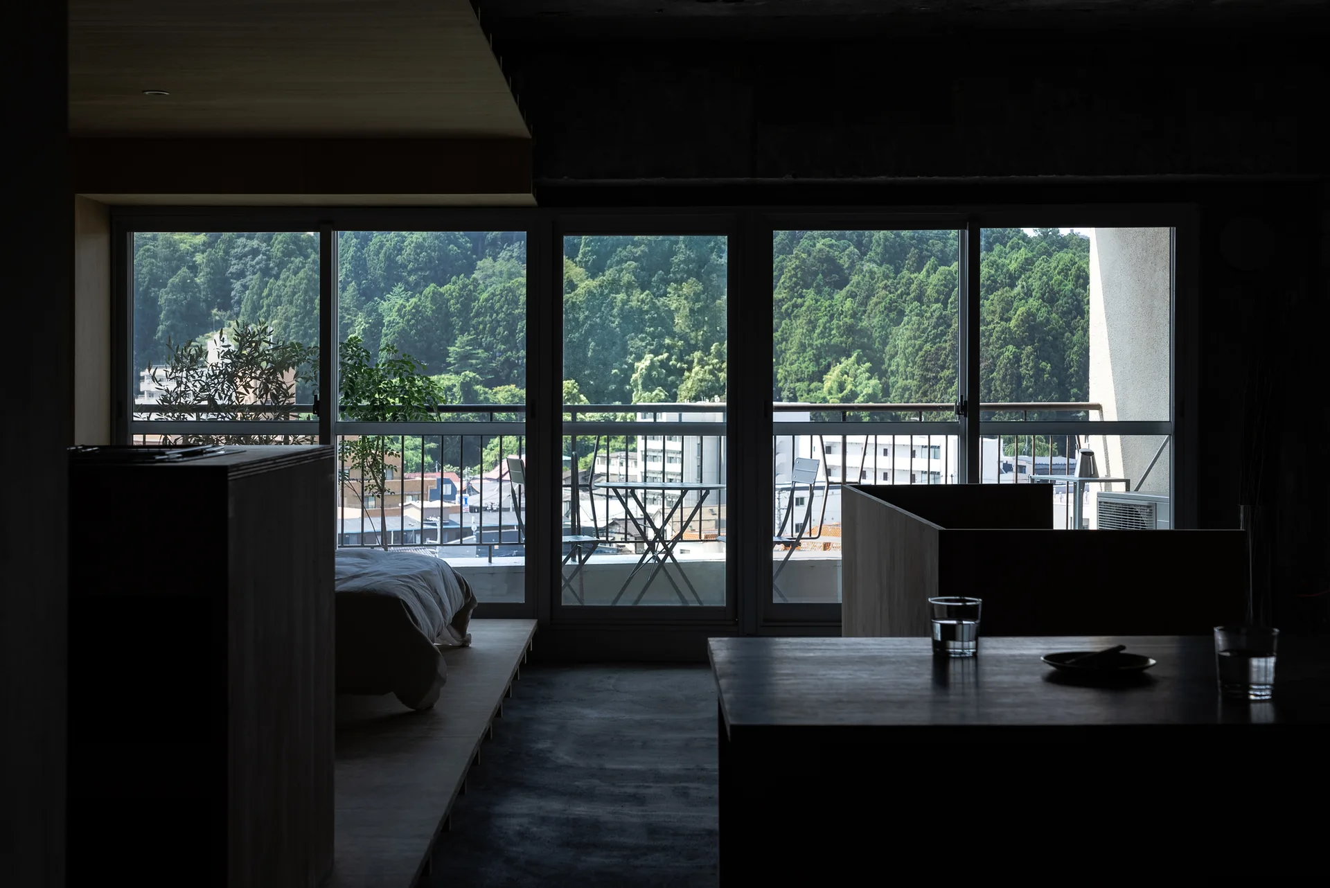 A dark minimalist bedroom features a low wooden platform bed beside large sliding glass windows looking out onto a balcony with a forest background.