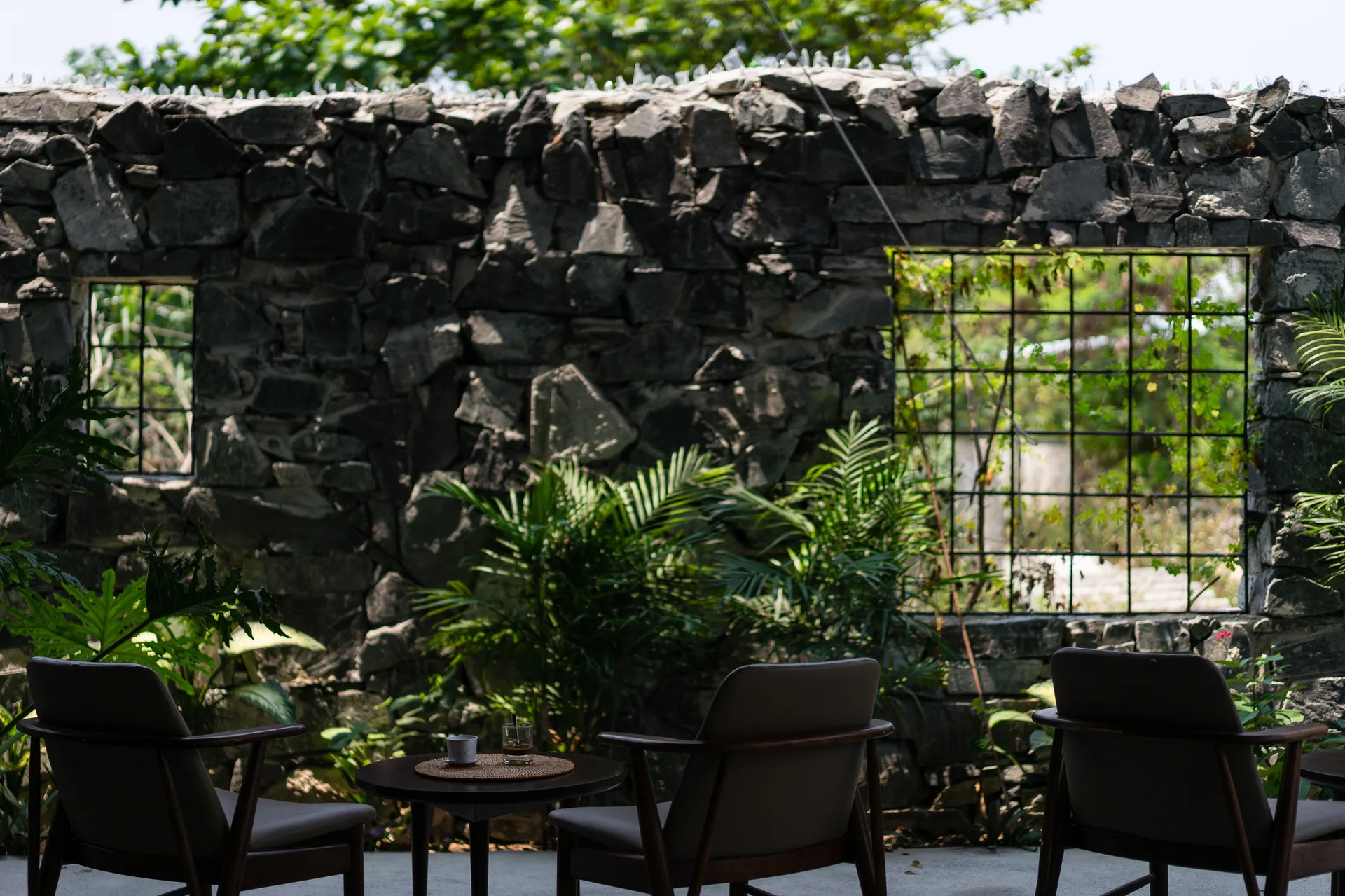 Three dark wood armchairs sit on a concrete patio in front of a rustic stacked stone wall with iron window grates and green plants.