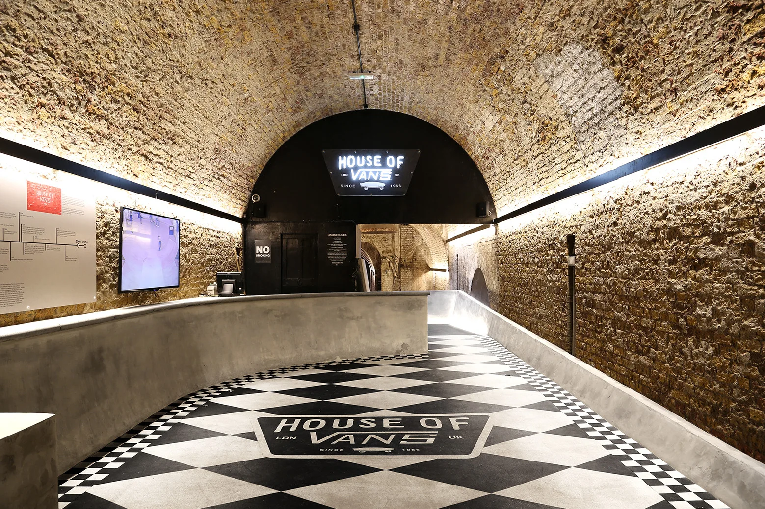 A long arched corridor features exposed brick barrel vaulted ceilings, a concrete reception counter, and a black and white checkered floor.