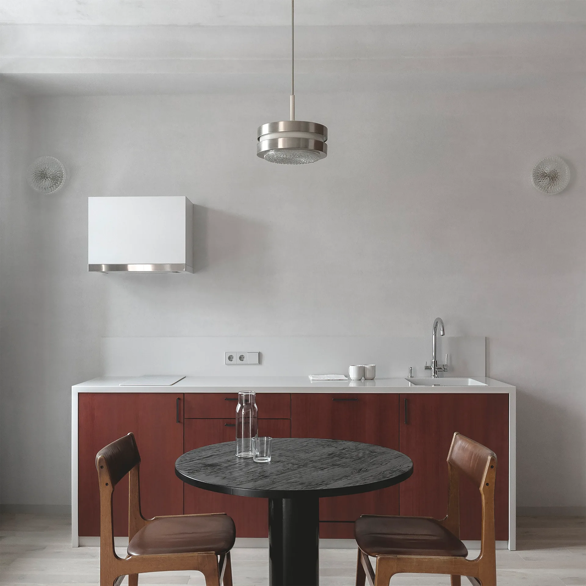 A minimalist kitchen features red-brown lower cabinets under a white countertop, paired with a round black dining table and two wooden chairs.