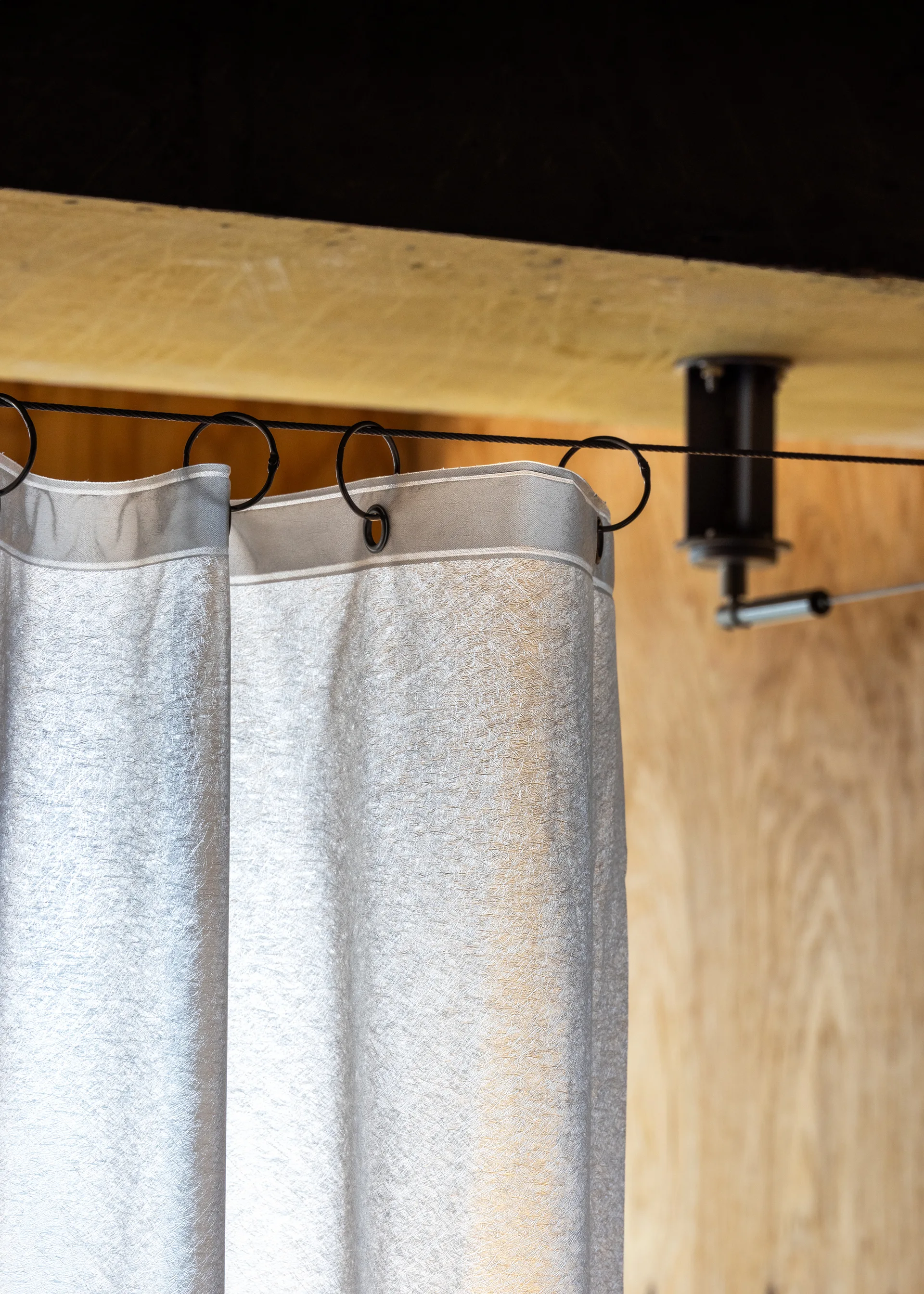 A close-up of a sheer grey curtain suspended by black rings from a thin metal tension wire against a light wood-grained wall panel.