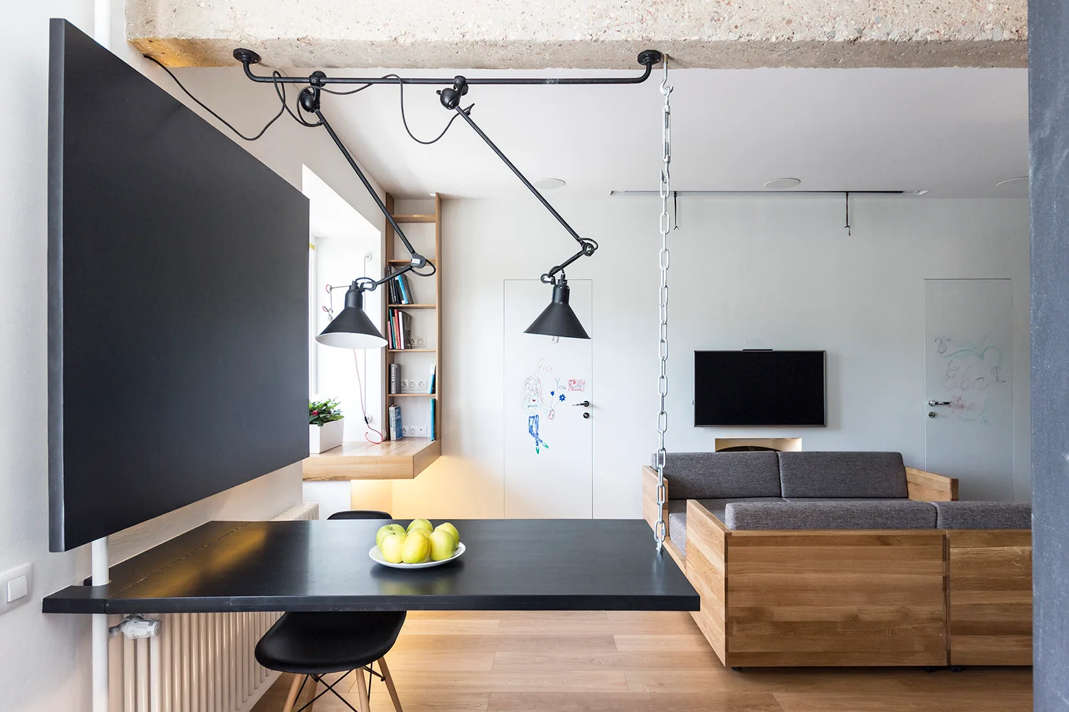 A black dining table suspended by a metal chain sits near a wood-framed grey sofa in a minimalist living room with an exposed concrete ceiling beam.