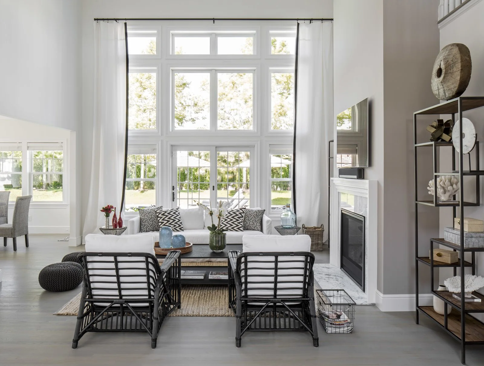 A bright, high-ceilinged living room features a symmetrical seating arrangement with black rattan armchairs, a white sofa, and a marble-surround fireplace.