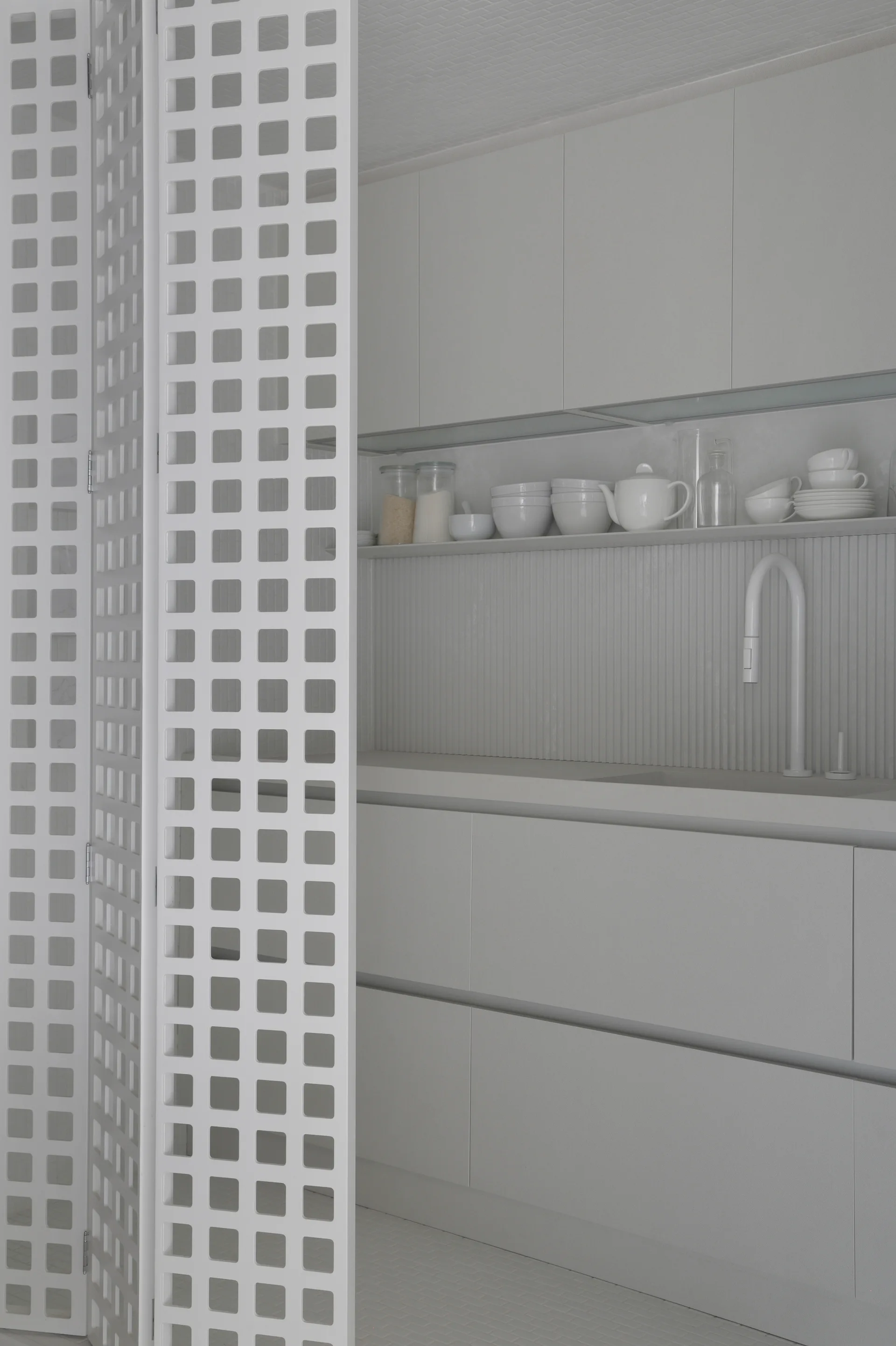 A minimalist all-white kitchen featuring matte flat-front cabinets, a grooved backsplash, a modern curved faucet, and a folding white grid screen divider.