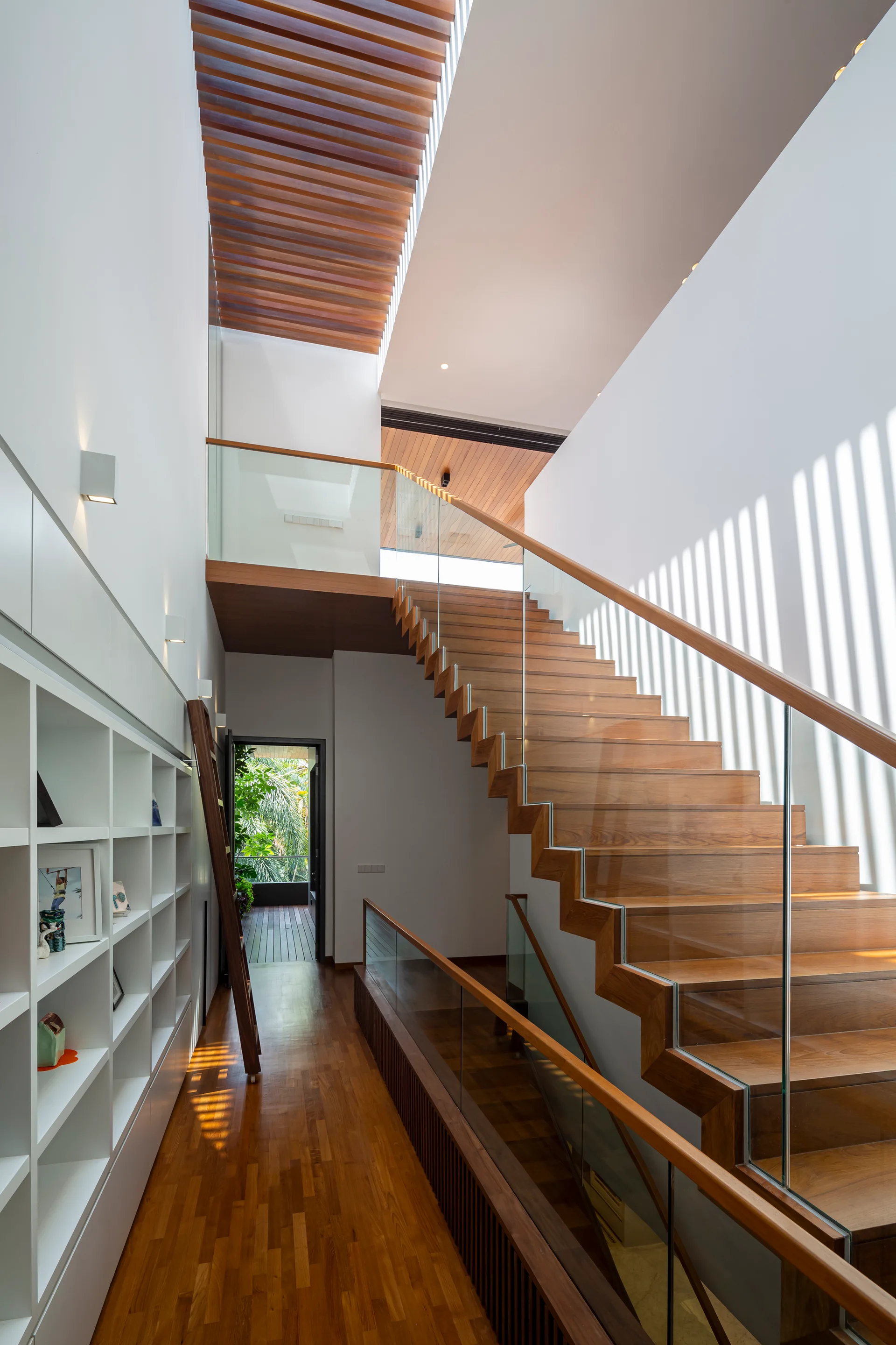 A modern wooden staircase with glass railings rises alongside a white wall cast with slatted linear shadows under a wood-slatted ceiling.