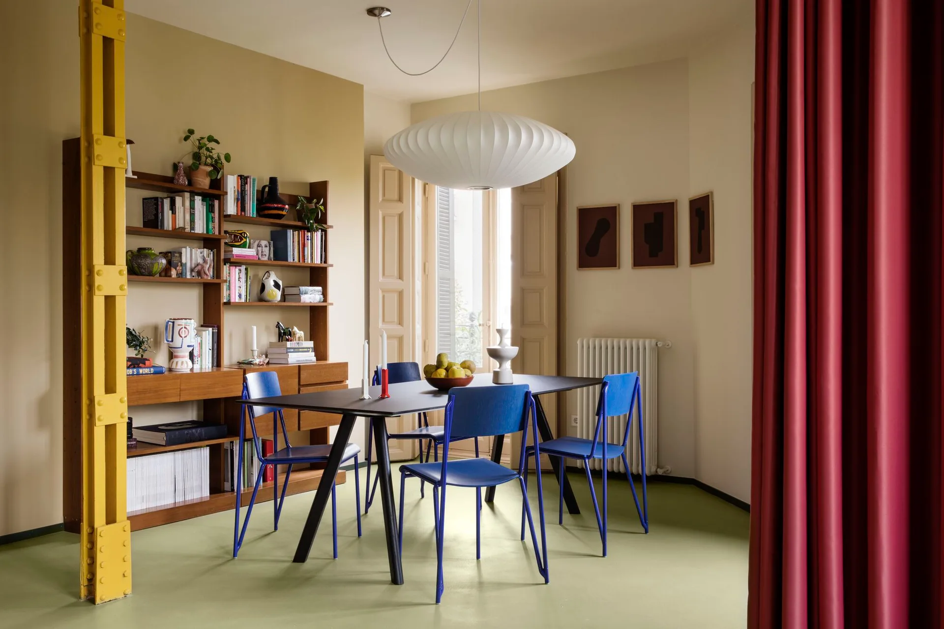 A dining room features cobalt blue chairs around a black table, a large white pendant light, a yellow steel beam, and a wooden shelving unit.