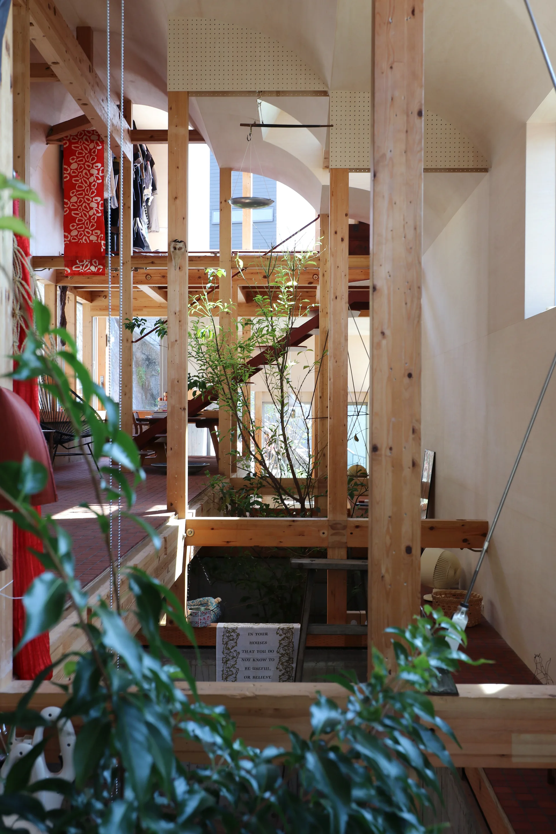 An open-concept multi-level interior features vertical wooden structural posts, a curved plywood vaulted ceiling, and live plants growing through the floor.