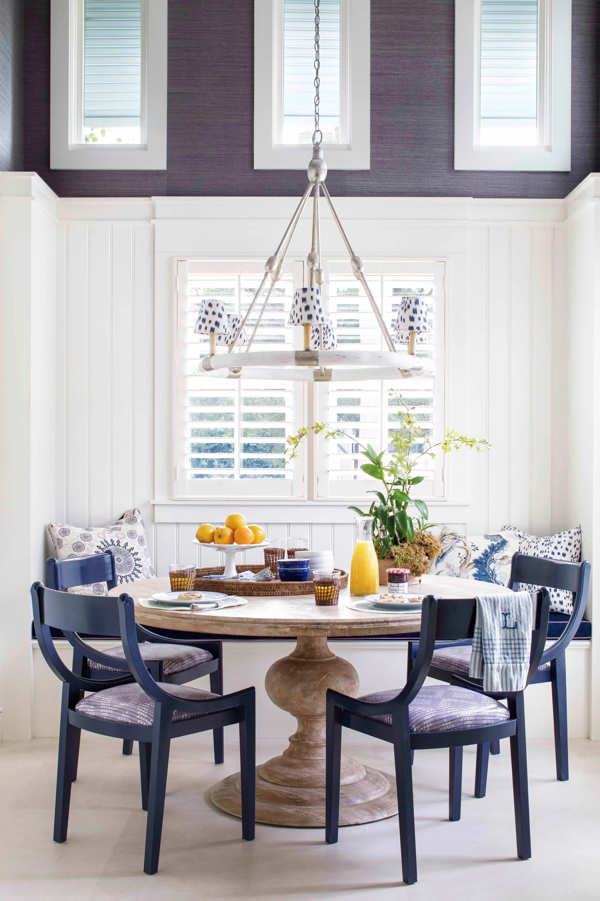 A bright dining nook features a round wooden pedestal table, navy chairs, built-in banquette seating, and windows with white plantation shutters.