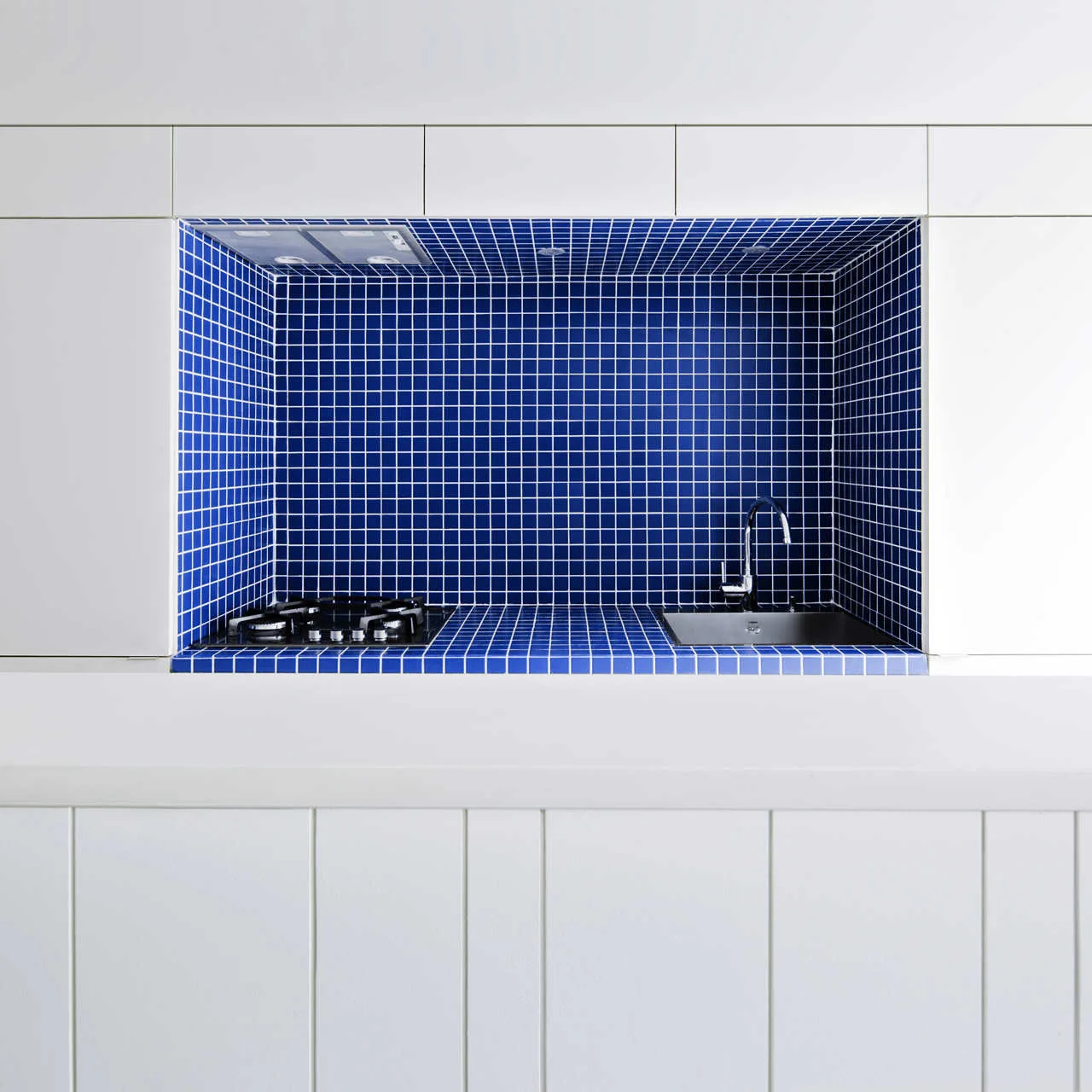 A recessed kitchen niche lined with small blue square tiles features a stainless steel sink, black gas stovetop, and white flat-front cabinetry.